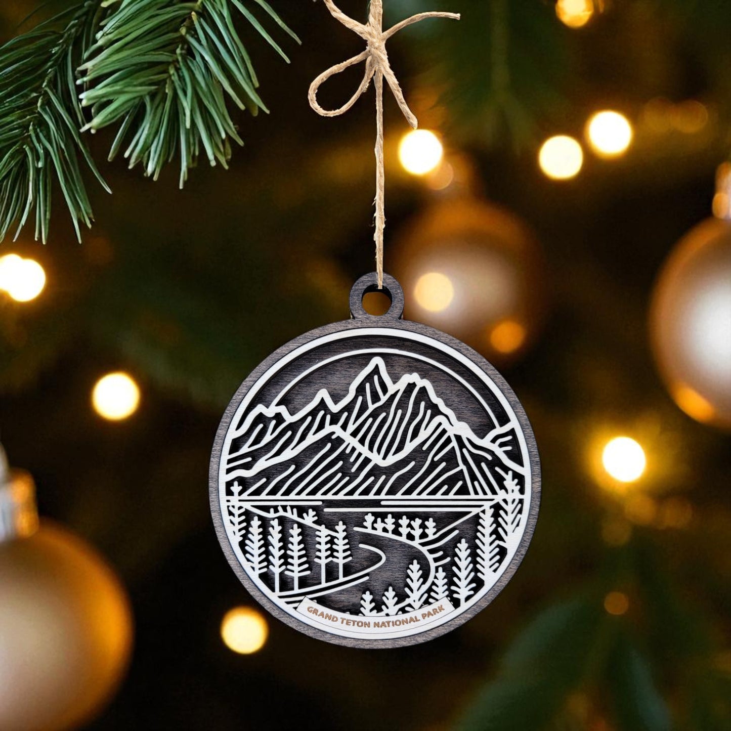 The Grand Teton National Park Ornament by Mirabilia Boutique—a wooden mountain landscape—hangs from a Christmas tree, with blurred lights and gold ornaments in the background.