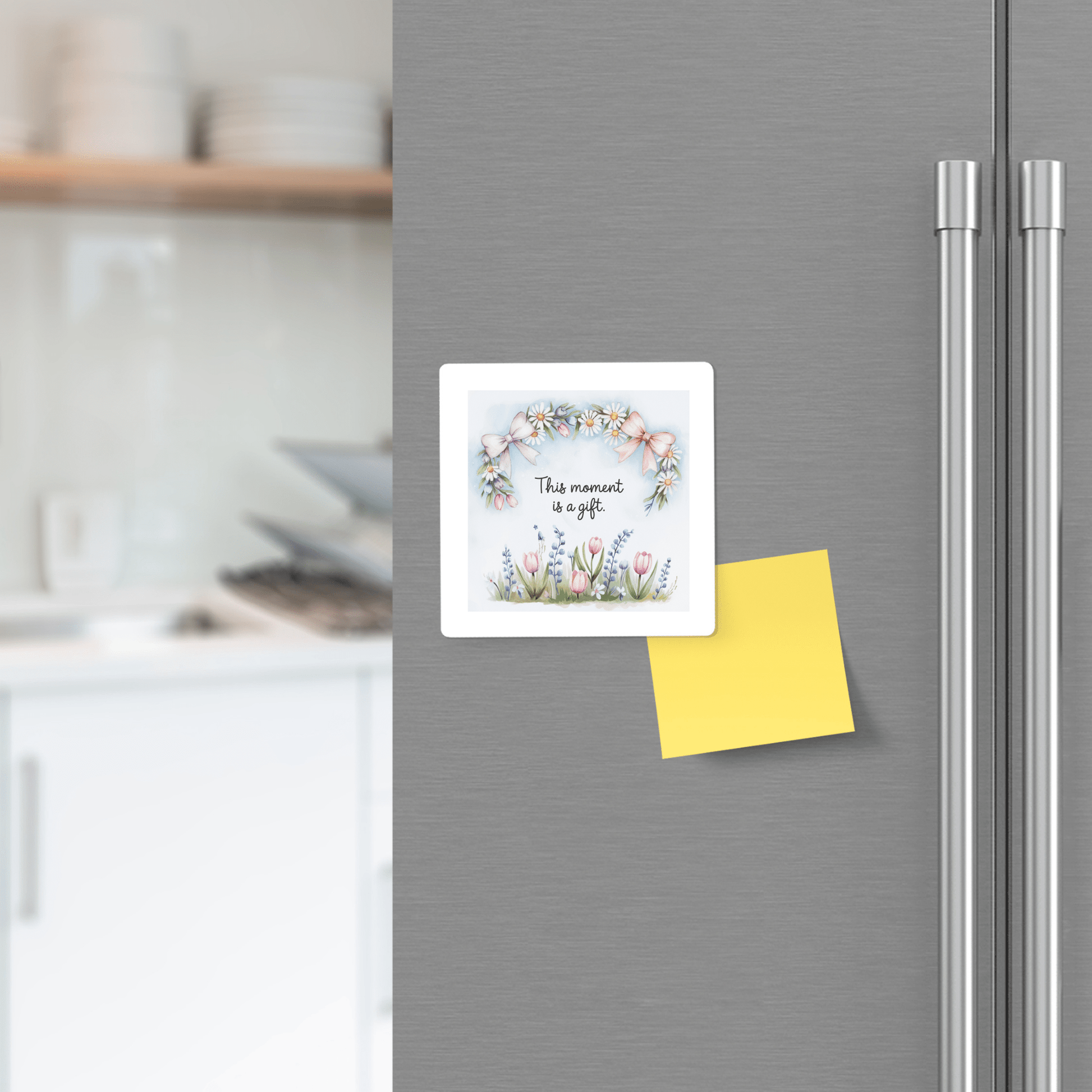 A Mirabilia Boutique "This Moment Is a Gift" floral spring quote magnet adds charm to a fridge door, which also features a yellow sticky note. A kitchen countertop and shelves are blurred in the background.