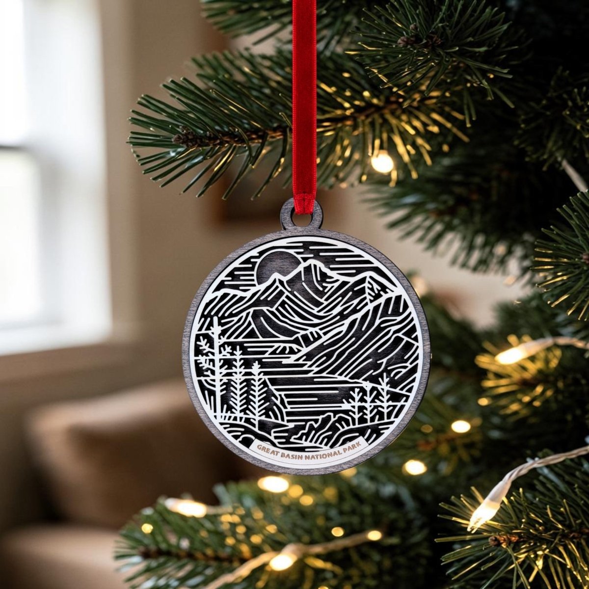A round Great Basin National Park Line Art Ornament by Mirabilia Boutique, featuring a detailed mountain and forest design, hangs from a red ribbon on a Christmas tree adorned with white string lights. The softly blurred background reveals part of a window and a couch.