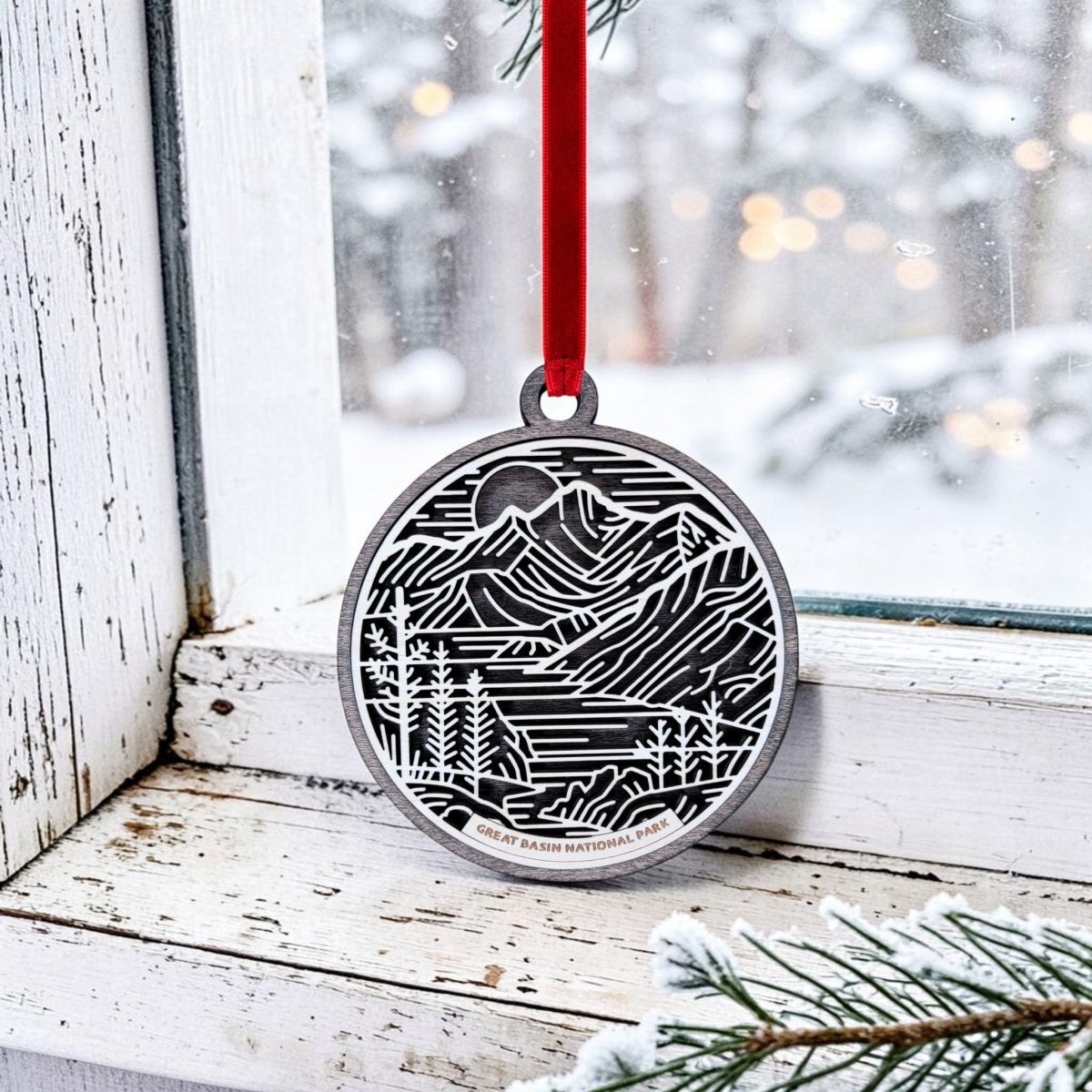 A Mirabilia Boutique Great Basin National Park Line Art Ornament, featuring an oval shape with a mountain and forest design, hangs from a red ribbon on a snowy window ledge. Frosted trees and snow can be seen outside.