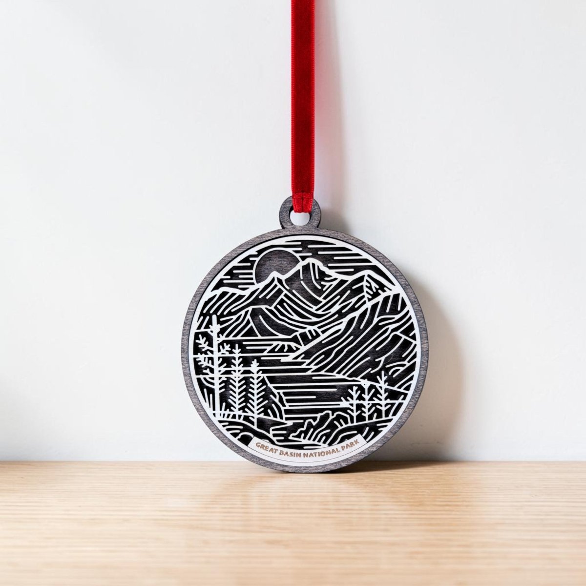 A round line art ornament from Mirabilia Boutique, featuring a black and white mountain landscape design inspired by Great Basin National Park, hangs from a red ribbon against a white wall and rests on a light wooden surface—an ideal addition to any wooden keepsake collection.