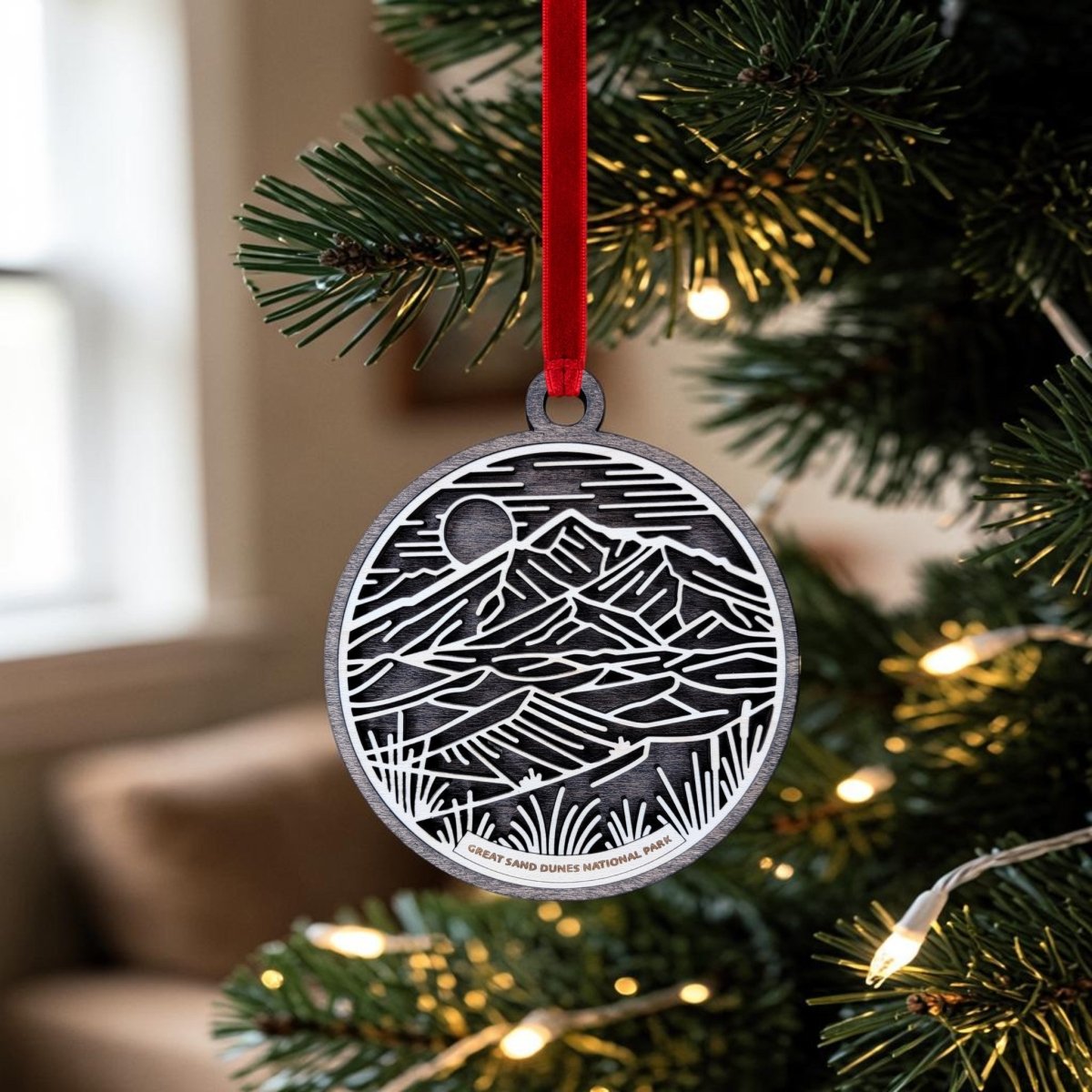 A metallic Great Sand Dunes National Park Line Art Ornament by Mirabilia Boutique, featuring an engraved mountain landscape and the inscription "Great Sand Dunes National Park," hangs from a Christmas tree branch by a red ribbon, surrounded by twinkling lights.