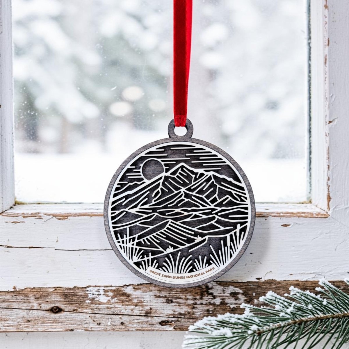 The Great Sand Dunes National Park Line Art Ornament by Mirabilia Boutique, featuring a mountain and grass design, hangs by a red ribbon in front of a snowy window. Frosted pine needles can be seen at the bottom right corner.