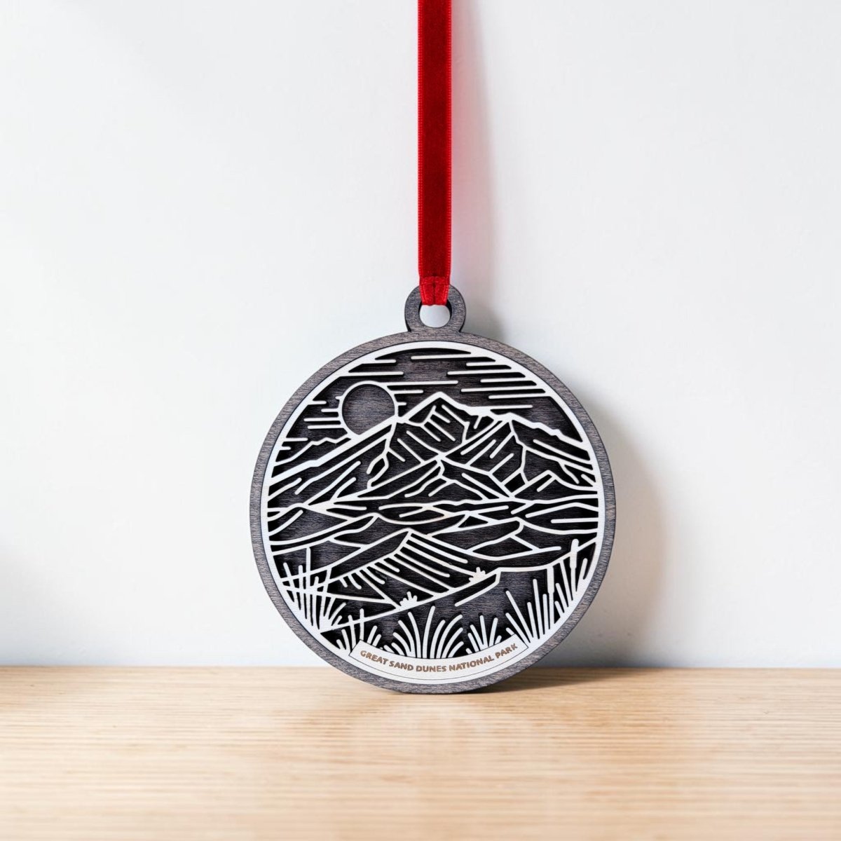 The Great Sand Dunes National Park Line Art Ornament by Mirabilia Boutique—a round wooden piece engraved with a landscape of mountains, sand dunes, and a sun—hangs from a red ribbon against a white wall, resting on a light wooden surface.