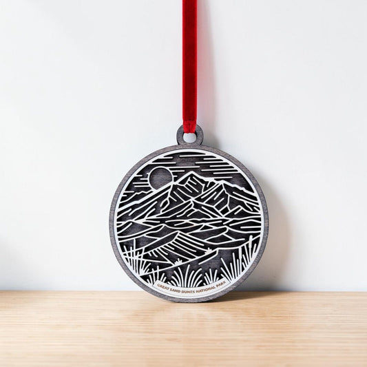 The Great Sand Dunes National Park Line Art Ornament by Mirabilia Boutique—a round wooden piece engraved with a landscape of mountains, sand dunes, and a sun—hangs from a red ribbon against a white wall, resting on a light wooden surface.