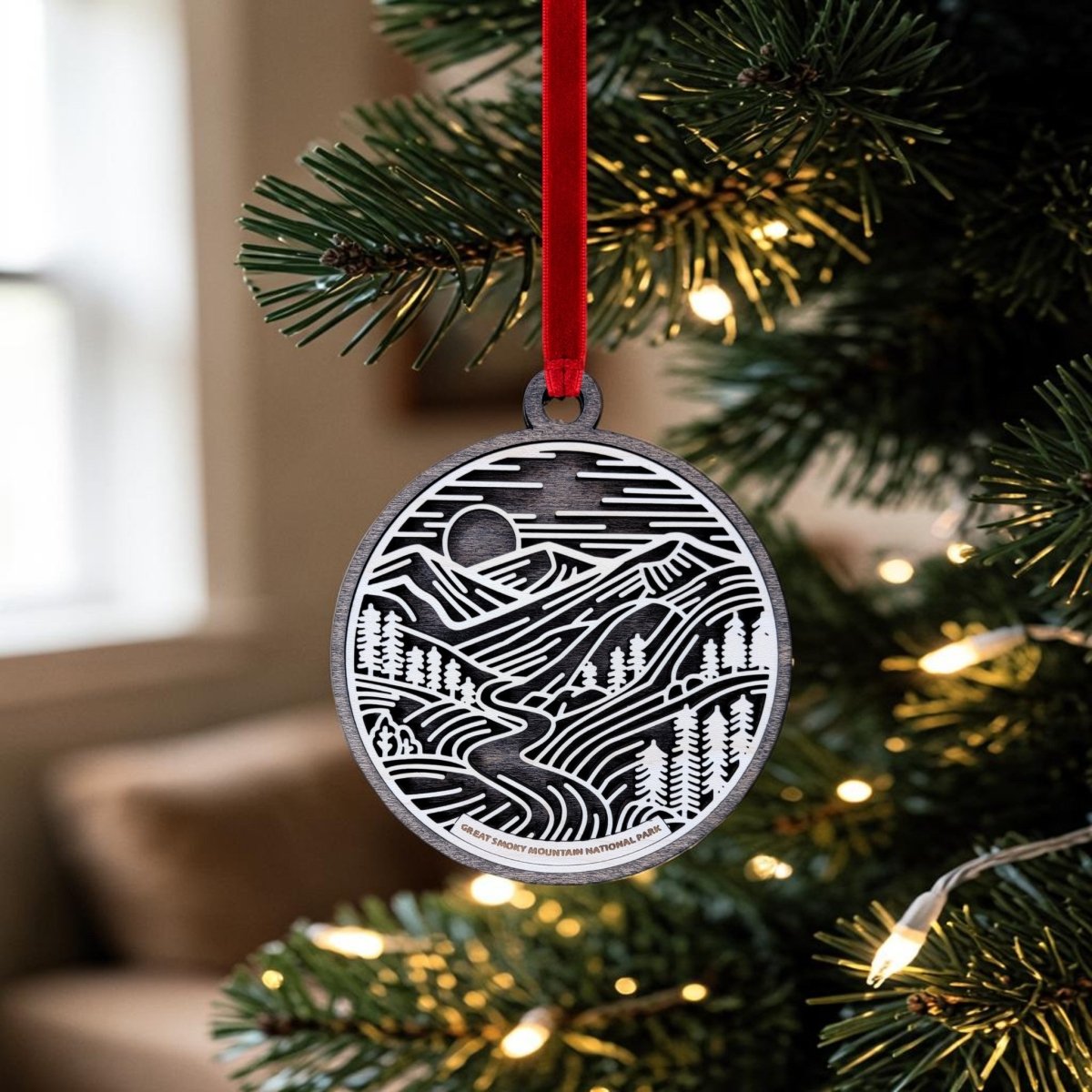 The Great Smoky Mountains National Park Line Art Ornament by Mirabilia Boutique features a black and white design of mountains, trees, and a river, hanging from a red ribbon on a lit Christmas tree—an ideal addition to any National Park keepsake collection.