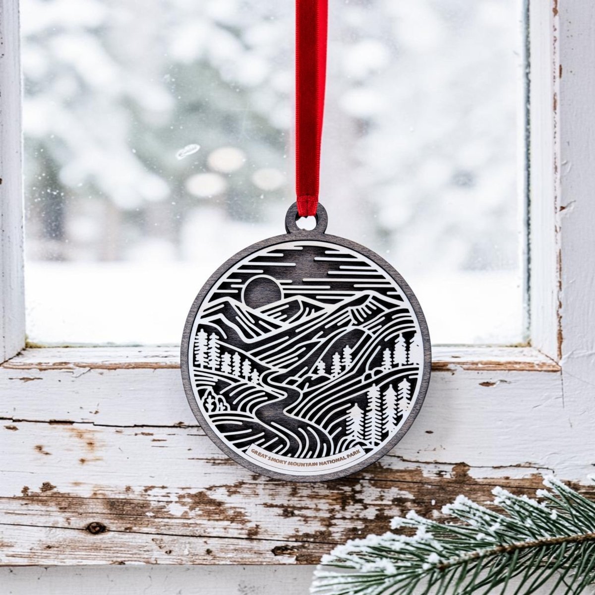 The Great Smoky Mountains National Park Line Art Ornament by Mirabilia Boutique, featuring an oval mountain and forest scene, hangs from a red ribbon on an old white window frame. Outside, snow and pine branches enhance the scene, making this National Park keepsake a charming winter display.