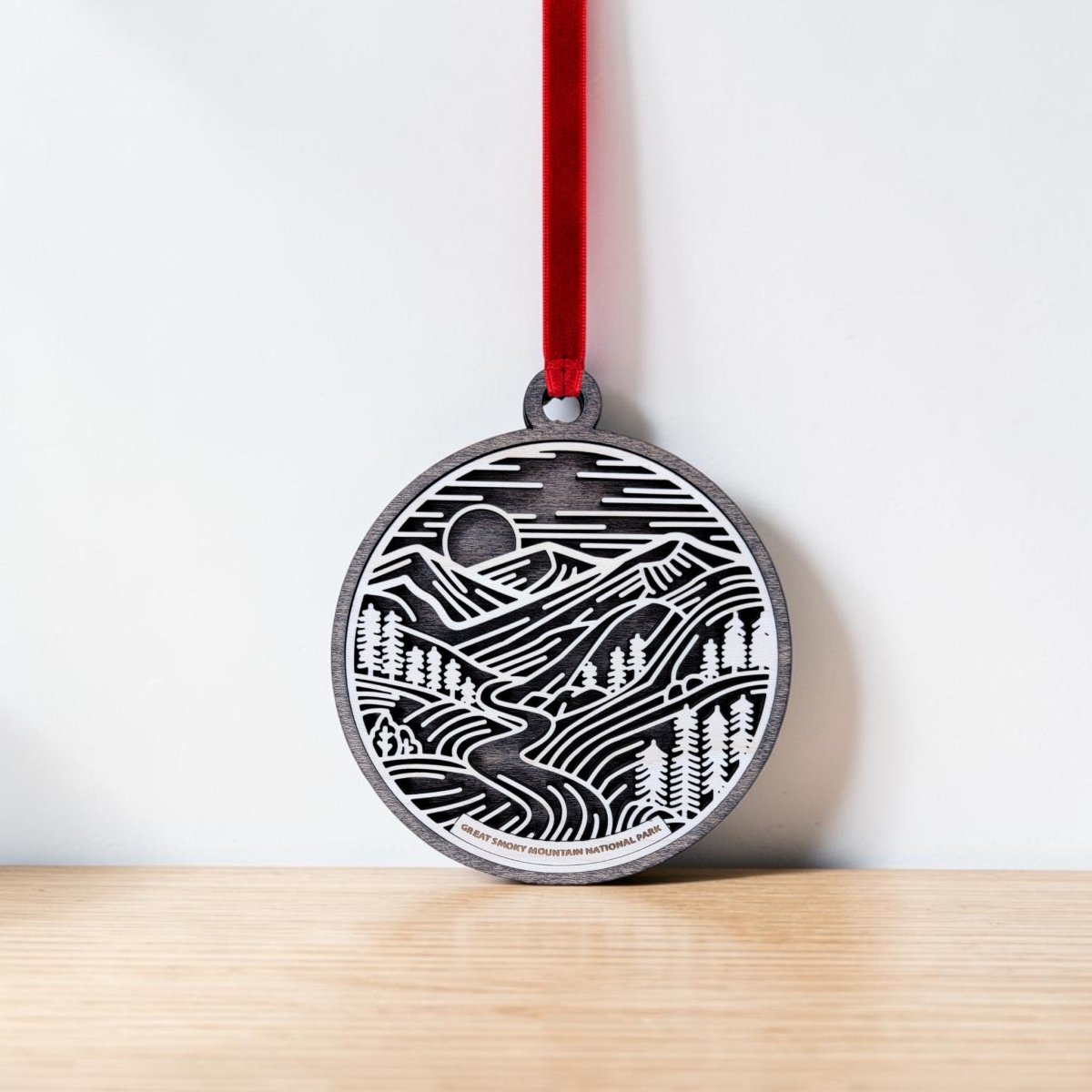 Introducing the Great Smoky Mountains National Park Line Art Ornament from Mirabilia Boutique—an elegant oval ornament with a red ribbon, showcasing a black and white engraved mountain landscape complete with trees, a river, and a sun or moon in the sky. Resting gracefully against a white wall on a wooden surface, this stunning line art piece makes an ideal National Park keepsake or Great Smoky Mountains ornament, capturing the serene beauty of nature in exquisite detail.