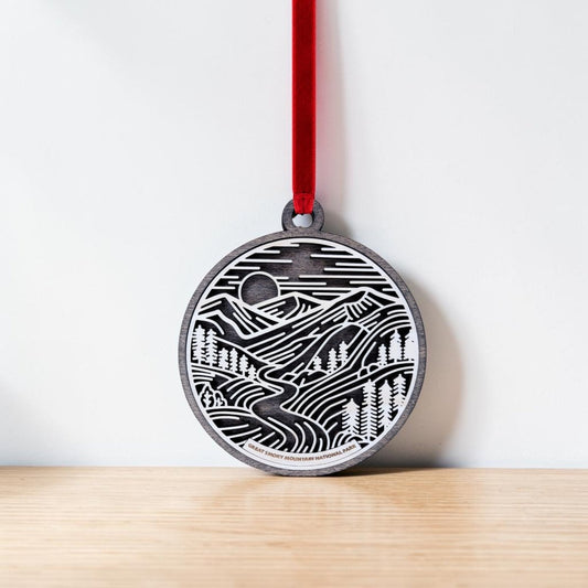 Introducing the Great Smoky Mountains National Park Line Art Ornament from Mirabilia Boutique—an elegant oval ornament with a red ribbon, showcasing a black and white engraved mountain landscape complete with trees, a river, and a sun or moon in the sky. Resting gracefully against a white wall on a wooden surface, this stunning line art piece makes an ideal National Park keepsake or Great Smoky Mountains ornament, capturing the serene beauty of nature in exquisite detail.