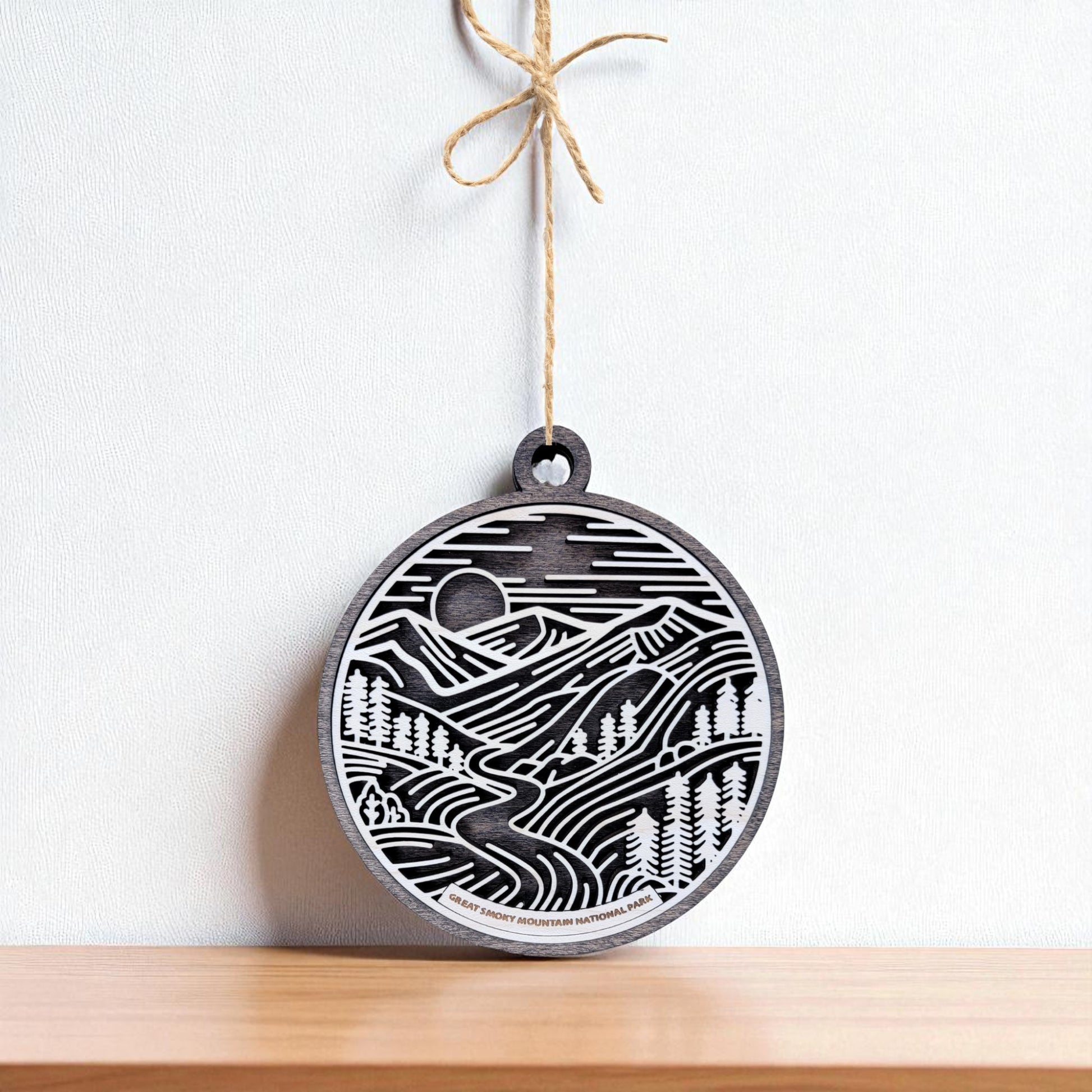 The Great Smoky Mountains Ornament by Mirabilia Boutique is a round, handcrafted wooden decoration featuring a mountain, trees, river, and sun design—perfect as a national park gift. Hangs on twine above a shelf against a white wall.