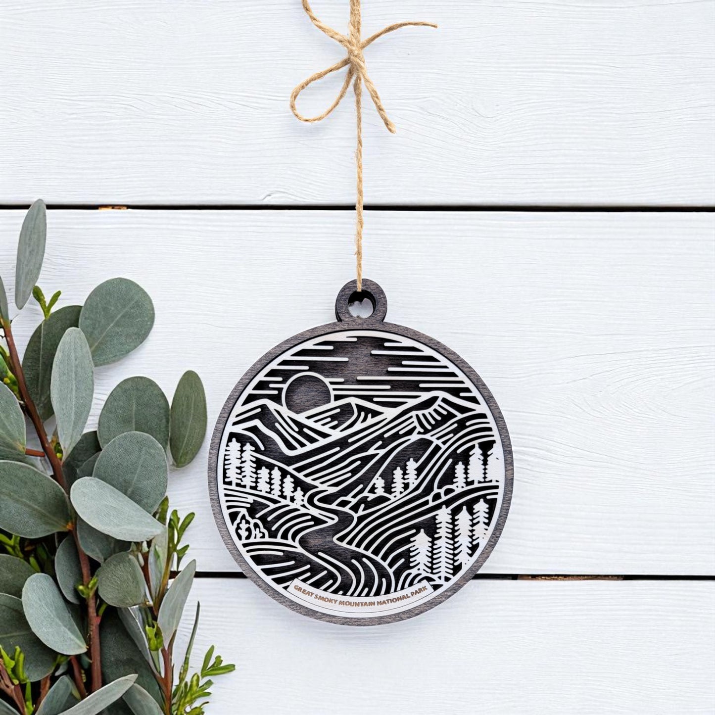 The Great Smoky Mountains Ornament by Mirabilia Boutique is a handcrafted round wood ornament featuring a black and white mountain design, hanging by twine on a white surface with green leafy branches to the left.