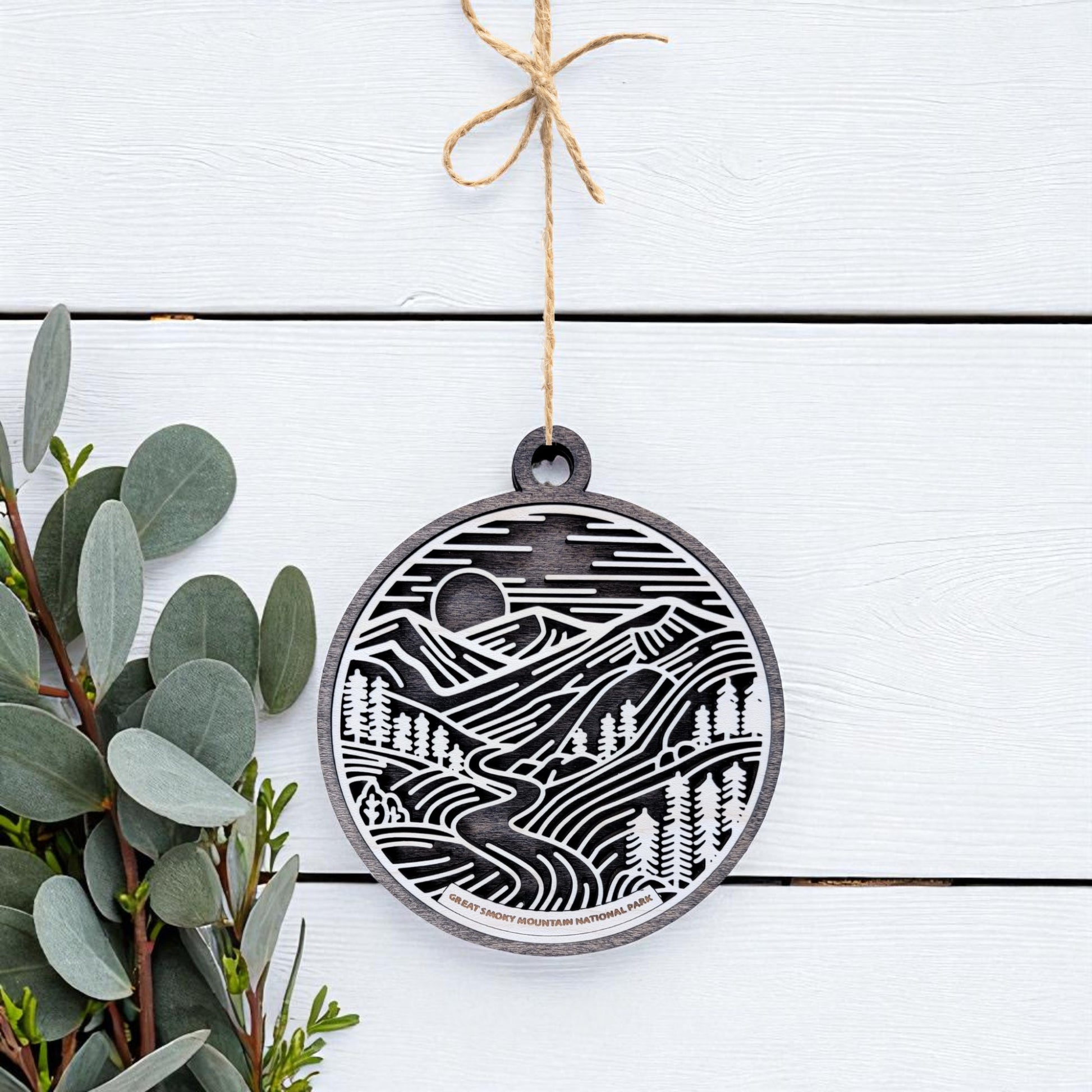 The Great Smoky Mountains Ornament by Mirabilia Boutique is a handcrafted round wood ornament featuring a black and white mountain design, hanging by twine on a white surface with green leafy branches to the left.