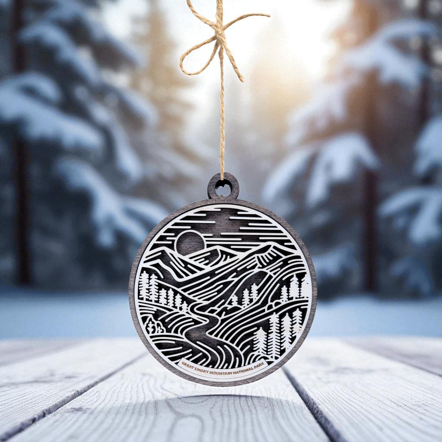 The Great Smoky Mountains Ornament by Mirabilia Boutique is a handcrafted wooden national park gift, featuring a black and white engraved mountain landscape and hanging by twine on a snowy wooden surface.