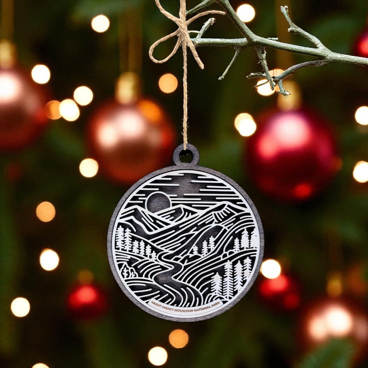 A round, black and white Great Smoky Mountains Ornament | Wooden National Park Gift by Mirabilia Boutique hangs from a tree, with red ornaments and warm lights softly illuminating the festive green branches.