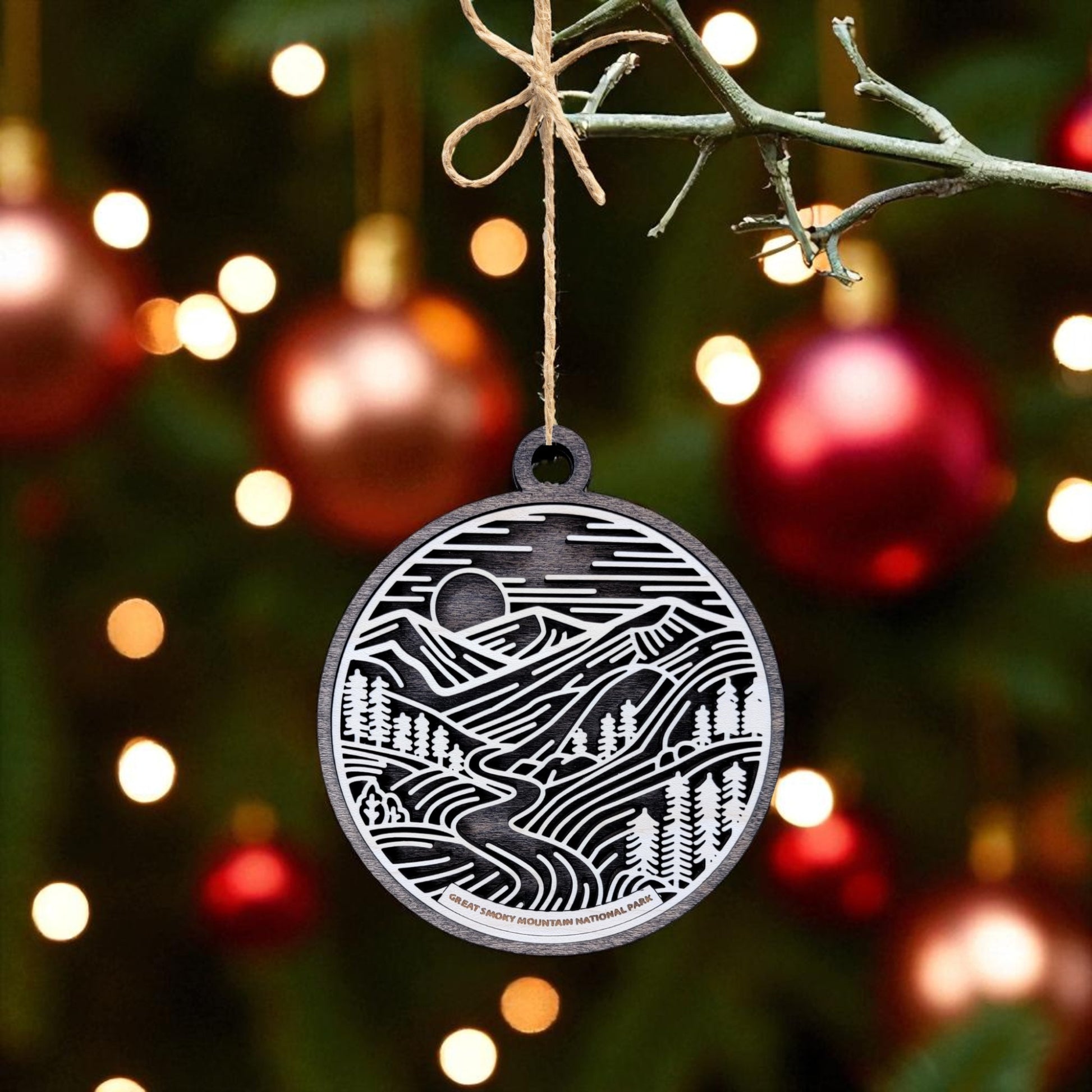 A round, black and white Great Smoky Mountains Ornament | Wooden National Park Gift by Mirabilia Boutique hangs from a tree, with red ornaments and warm lights softly illuminating the festive green branches.