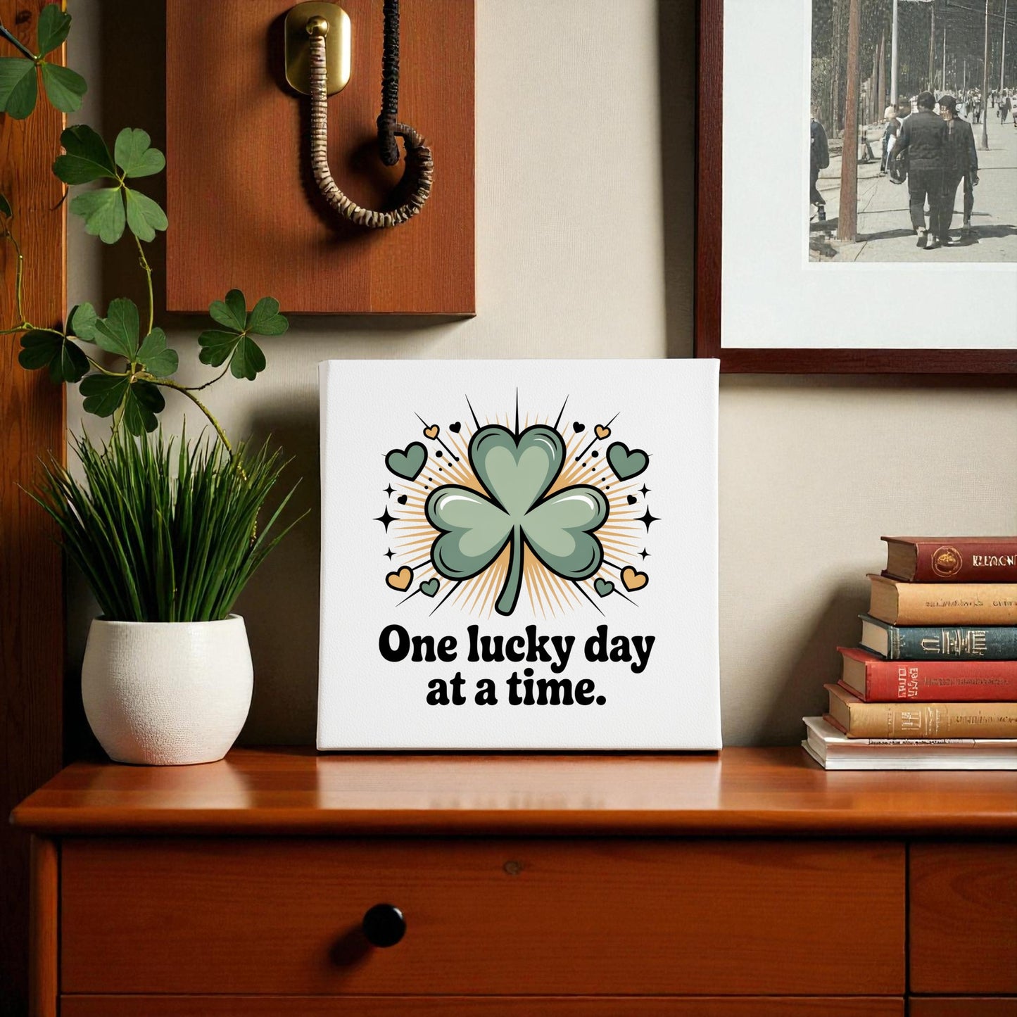 The Mirabilia Boutique "One Lucky Day at a Time" canvas brings optimism to your home decor, displayed on a wooden table next to a potted plant, vintage phone, and photo.