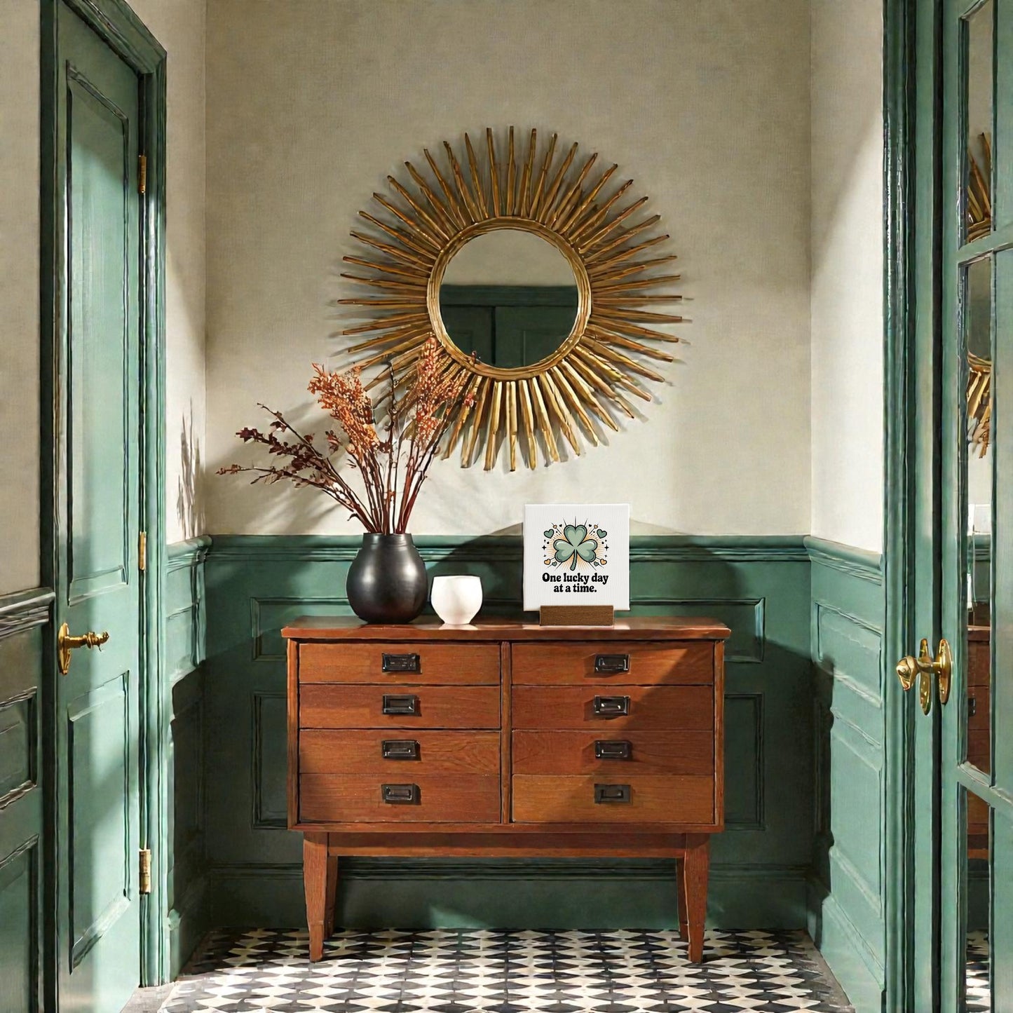 A wooden sideboard with drawers stands against green paneled walls and a checkered tile floor. Atop it sits black and white vases, a card, and Mirabilia Boutique’s “One Lucky Day at a Time” 4x4 mini sign—an ideal encouragement gift. A round sunburst mirror hangs above.