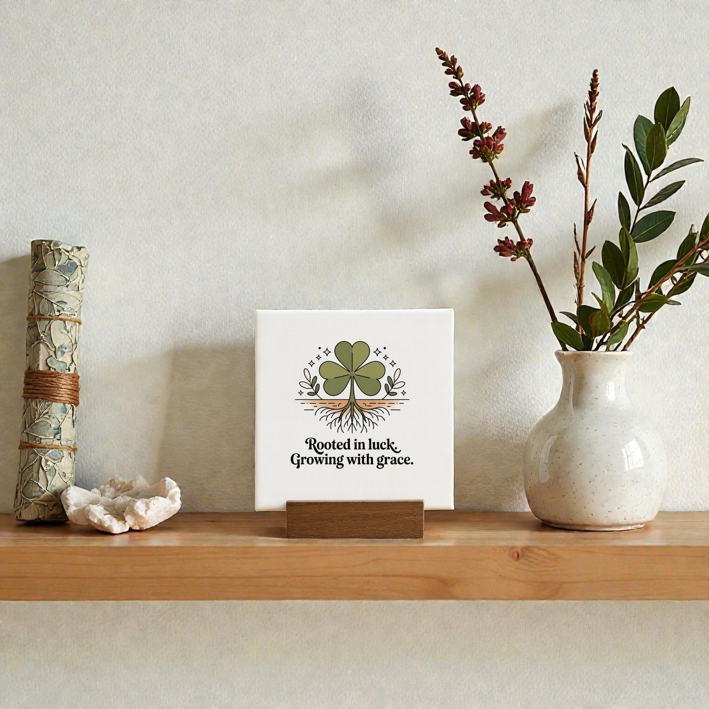 A wooden shelf showcases a scroll, a crystal, mini botanical decor, and the Mirabilia Boutique "Rooted in Luck, Growing with Grace" 4x4 canvas sign featuring a four-leaf clover, plus a ceramic vase with branches bearing small red leaves.