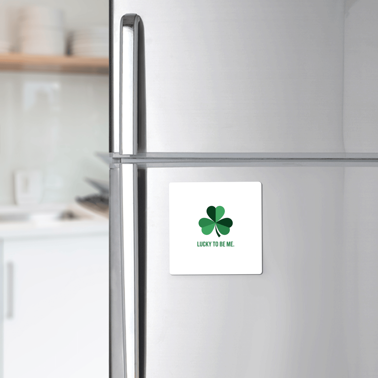 A stainless steel refrigerator features the Mirabilia Boutique Lucky to Be Me Shamrock Magnet—a white square magnet with a green four-leaf clover and "LUCKY TO BE ME" text—against a softly blurred kitchen background.