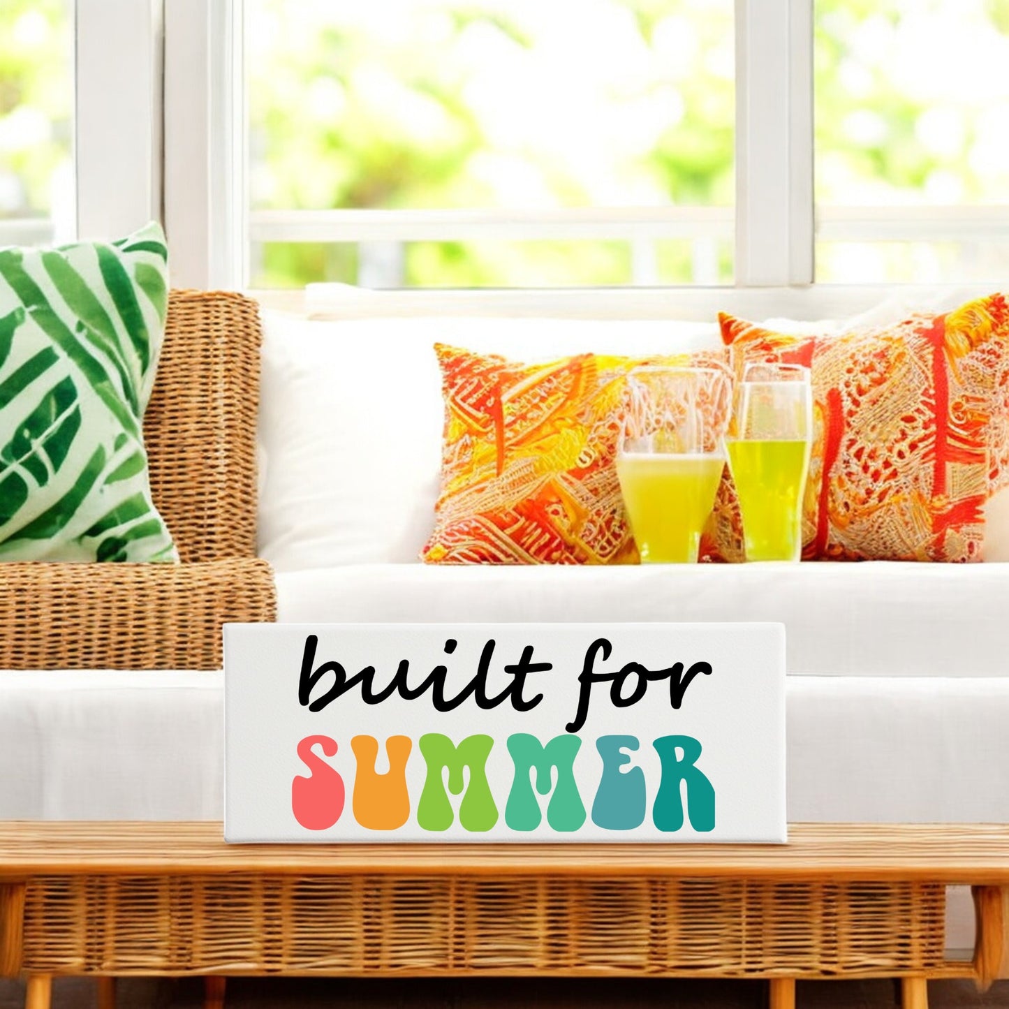 A wicker couch with colorful cushions faces a large window. On the table sits the Mirabilia Boutique "Built for Summer Canvas – Retro Shelf Sign," perfect for adding retro charm to any summer decor.