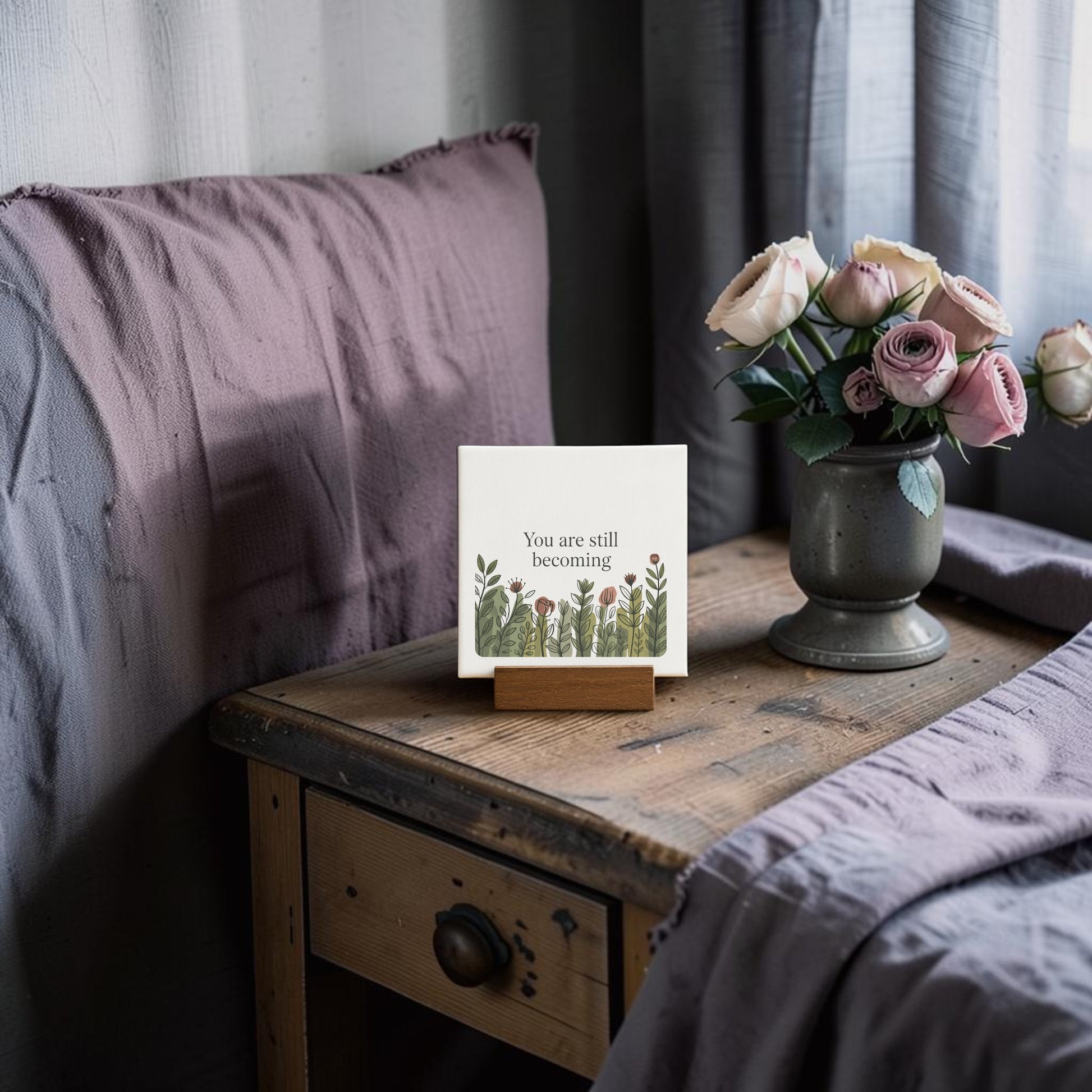 A rustic wooden stand displays the Mirabilia Boutique "You Are Still Becoming" Mini Canvas Sign with botanical art, set beside a vase of pink and white roses near a neatly made bed with lavender bedding.