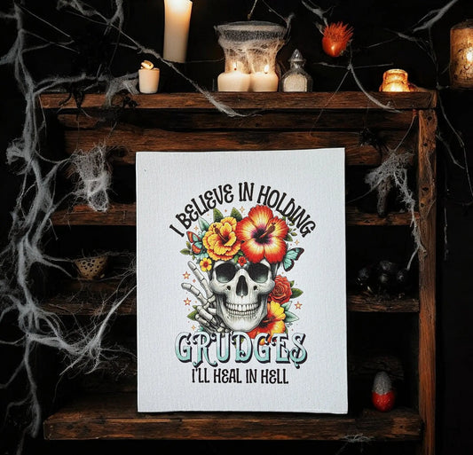 A captivating piece of wall art, the "I Believe in Holding Grudges Print" by Mirabilia Boutique, showcases a skull embellished with red and yellow flowers and green leaves. It includes the bold phrase: "I believe in holding grudges, I'll heal in hell." This striking print is ideal for edgy home decor and makes a statement when displayed on a wooden shelf surrounded by candles and cobwebs.