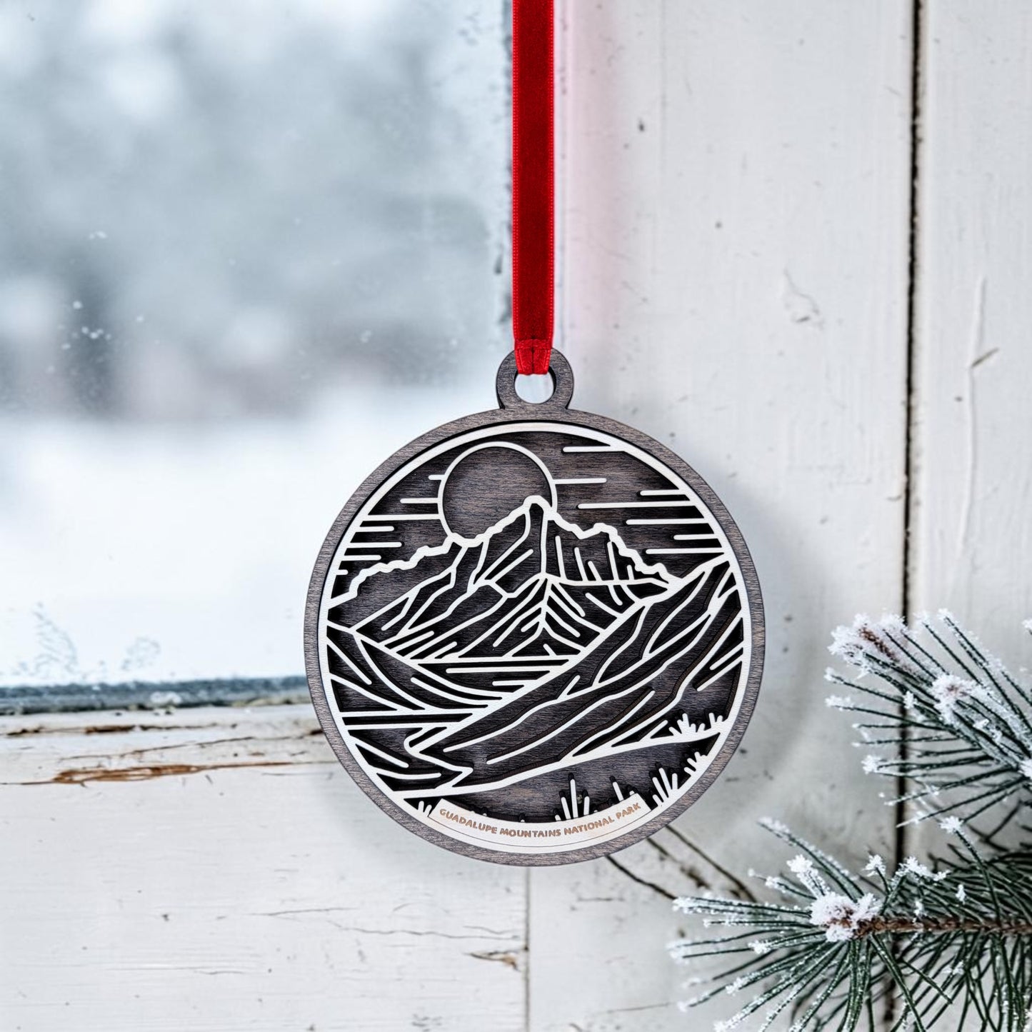The Guadalupe Mountains National Park Line Art Christmas Ornament by Mirabilia Boutique, featuring a metallic mountain and sun design, hangs from a red ribbon on a white window. Outside, snowy scenery can be seen, while frosted pine branches rest at the windowsill, capturing the peaceful essence of the park.