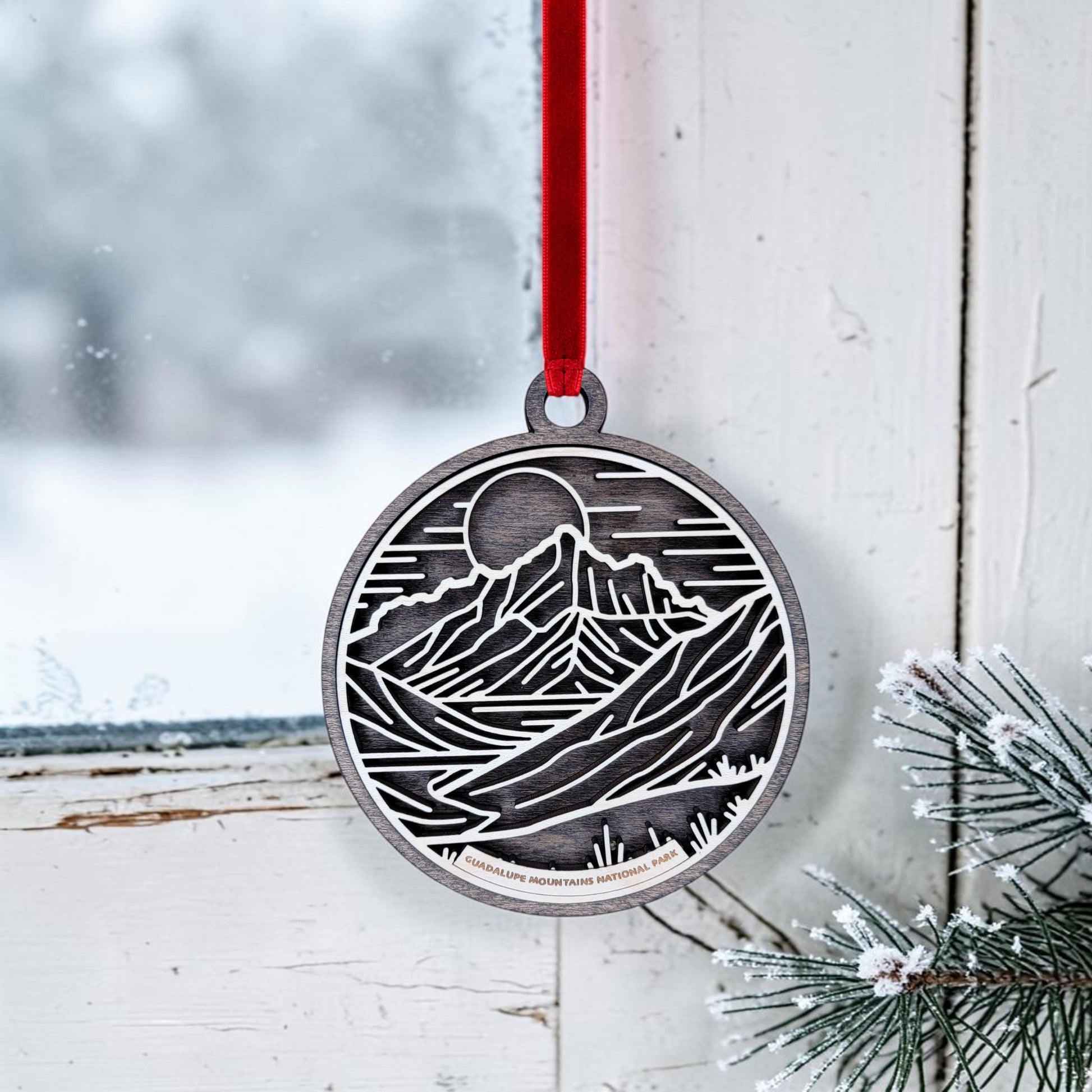 The Guadalupe Mountains National Park Line Art Christmas Ornament by Mirabilia Boutique, featuring a metallic mountain and sun design, hangs from a red ribbon on a white window. Outside, snowy scenery can be seen, while frosted pine branches rest at the windowsill, capturing the peaceful essence of the park.