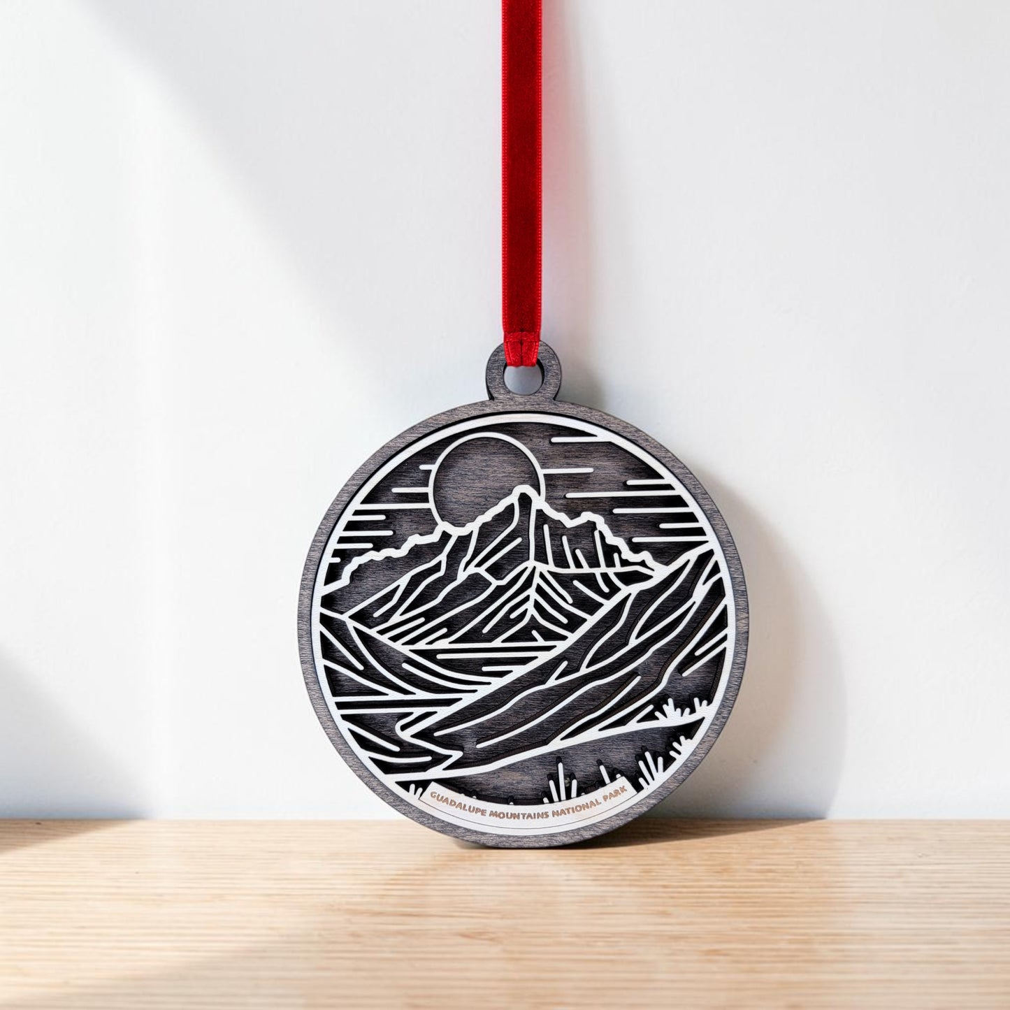 The Guadalupe Mountains National Park Line Art Christmas Ornament by Mirabilia Boutique features a round wooden design with a black and white mountain landscape, showcasing a sun or moon and river in line art style. It is displayed hanging by a red ribbon against a white background on a wooden surface.