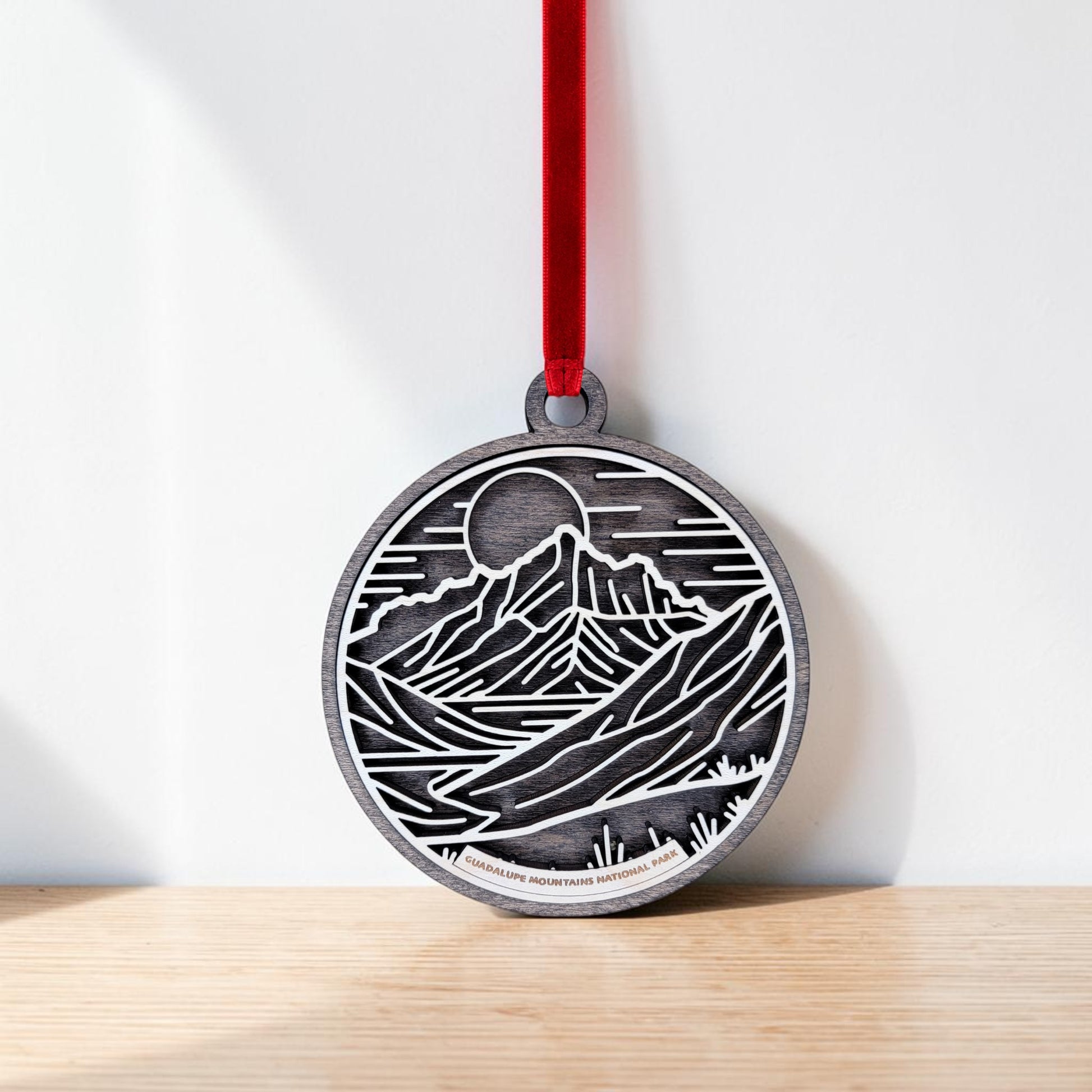 The Guadalupe Mountains National Park Line Art Christmas Ornament by Mirabilia Boutique features a round wooden design with a black and white mountain landscape, showcasing a sun or moon and river in line art style. It is displayed hanging by a red ribbon against a white background on a wooden surface.