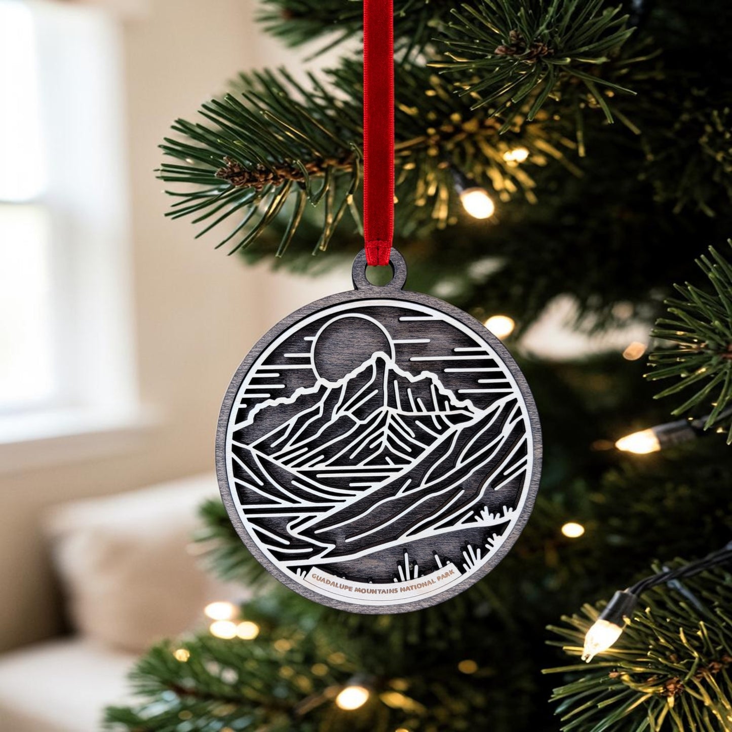 The Guadalupe Mountains National Park Line Art Christmas Ornament by Mirabilia Boutique, featuring a minimalist mountain landscape design, hangs from a red ribbon on a Christmas tree decorated with twinkling lights. In the softly blurred background, a window and couch add to the cozy holiday ambiance.
