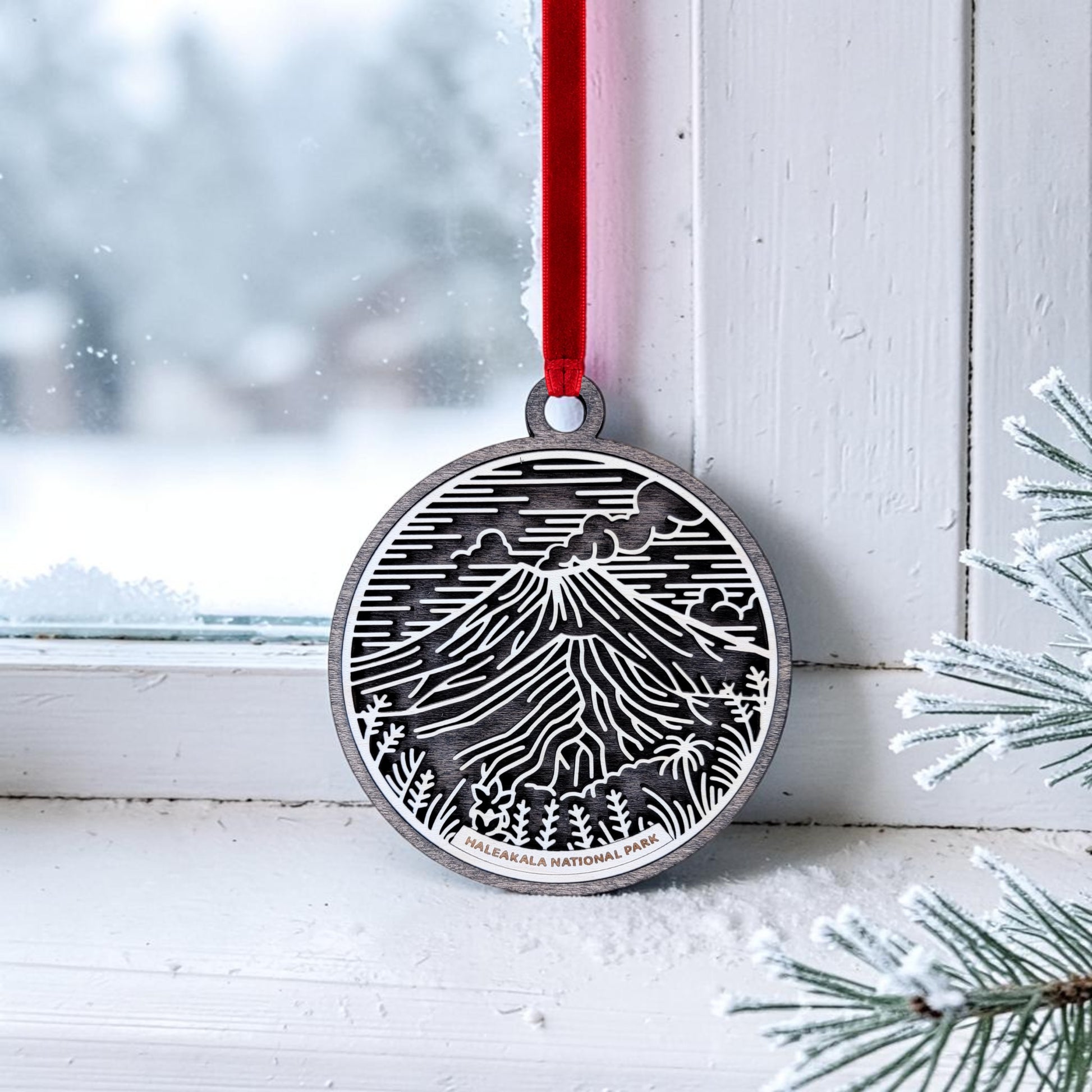 A metal ornament featuring a mountain and trees design hangs by a red ribbon on a snowy window, with pine needles in the corner—a beautiful Haleakalā National Park Line Art Christmas Ornament by Mirabilia Boutique, perfect for anyone who treasures keepsakes inspired by volcanic craters.