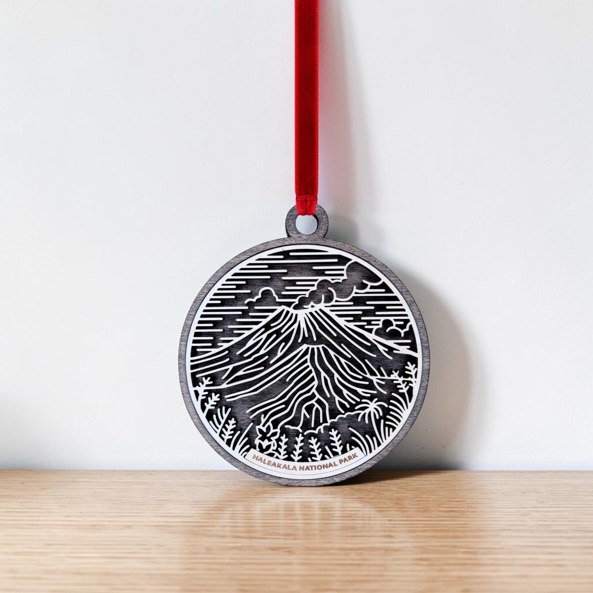 The Haleakalā National Park Line Art Christmas Ornament by Mirabilia Boutique is a round metal keepsake featuring a line art design of mountains and clouds inspired by the park’s famous volcanic crater. It hangs from a red ribbon against a white wall above a wooden surface.