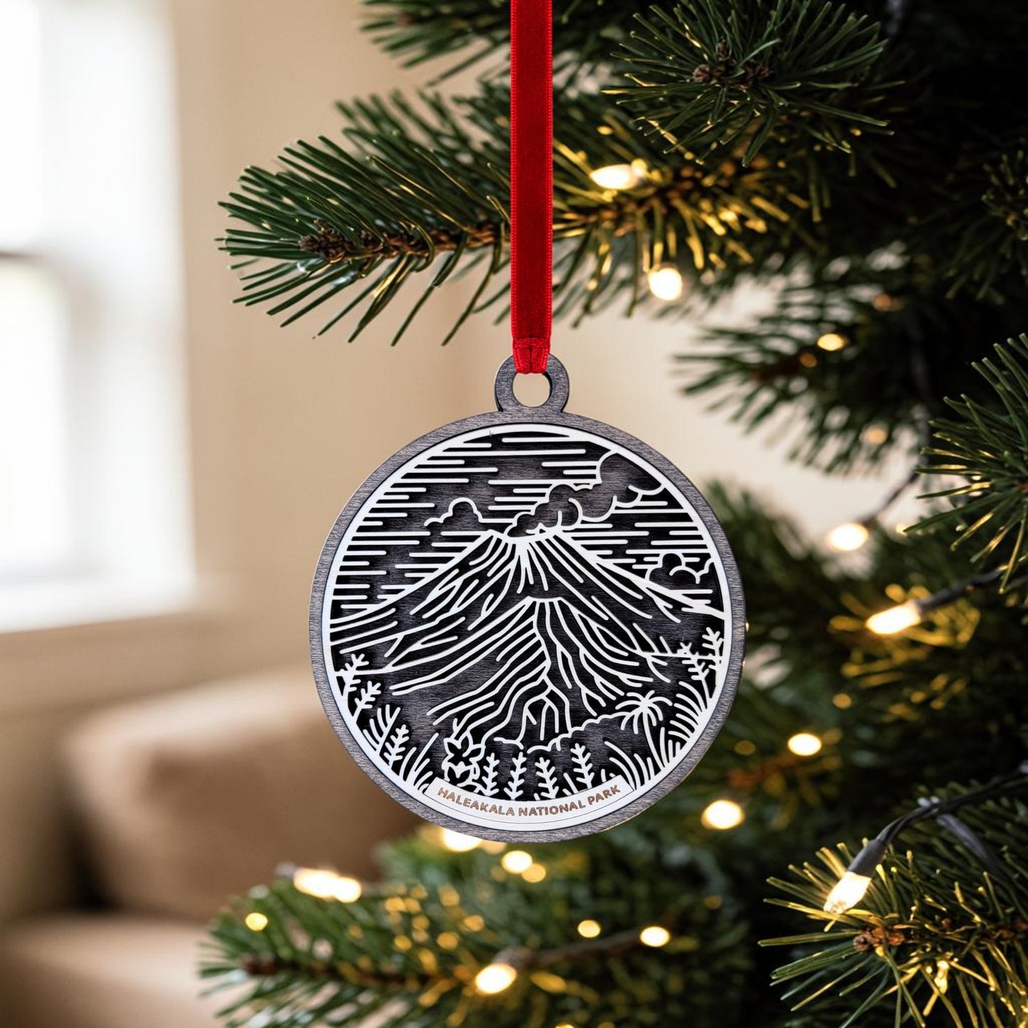 A metal Haleakalā National Park Line Art Christmas Ornament by Mirabilia Boutique, featuring a mountain scene and labeled “Haleakalā National Park,” hangs from a red ribbon on a lit Christmas tree—making it the perfect volcanic crater keepsake for your holiday décor.