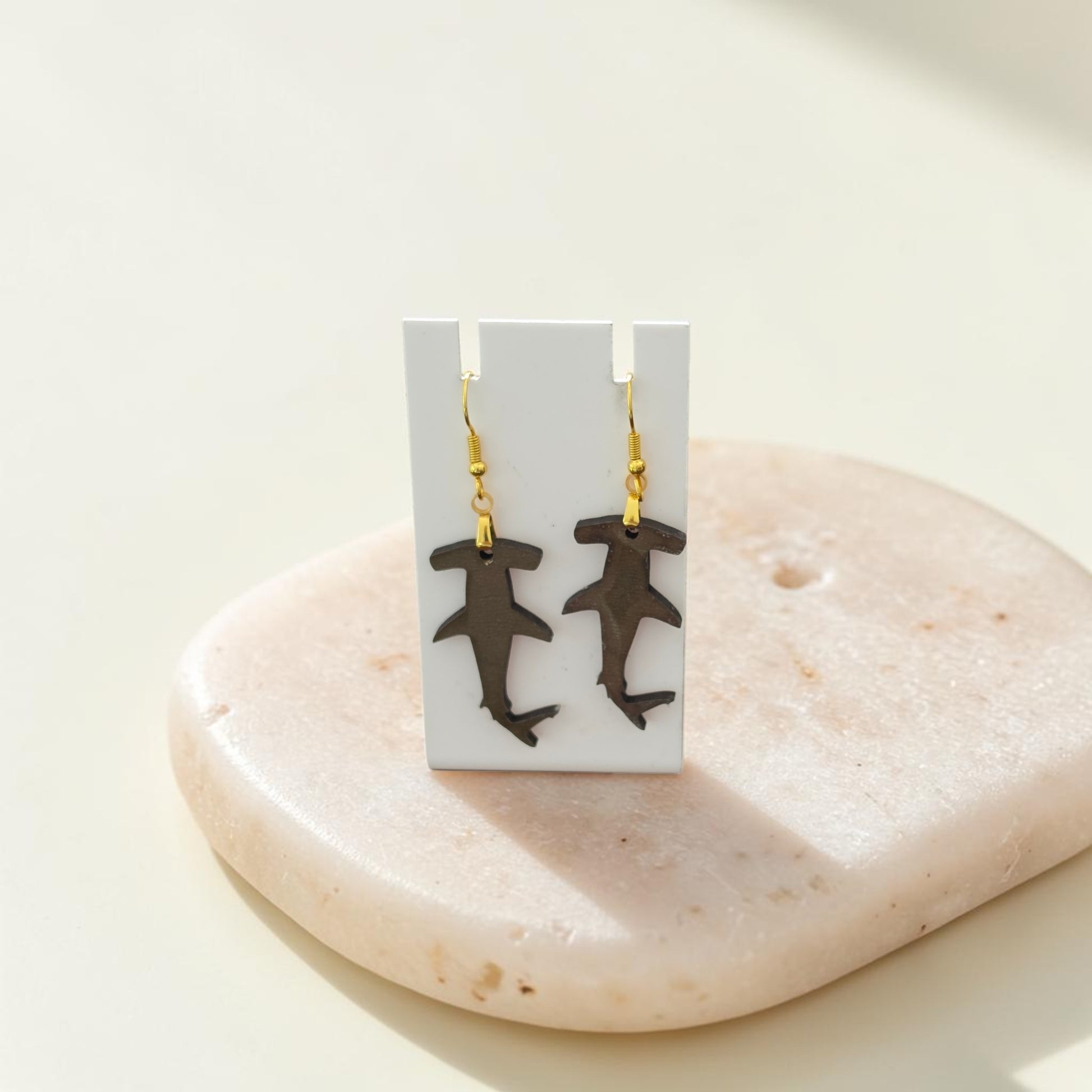 The Hammerhead Shark Earrings | Lasercut Walnut Wood by Mirabilia Boutique hang from a white display card with gold hooks, shown on a smooth, light-colored stone against a neutral backdrop.