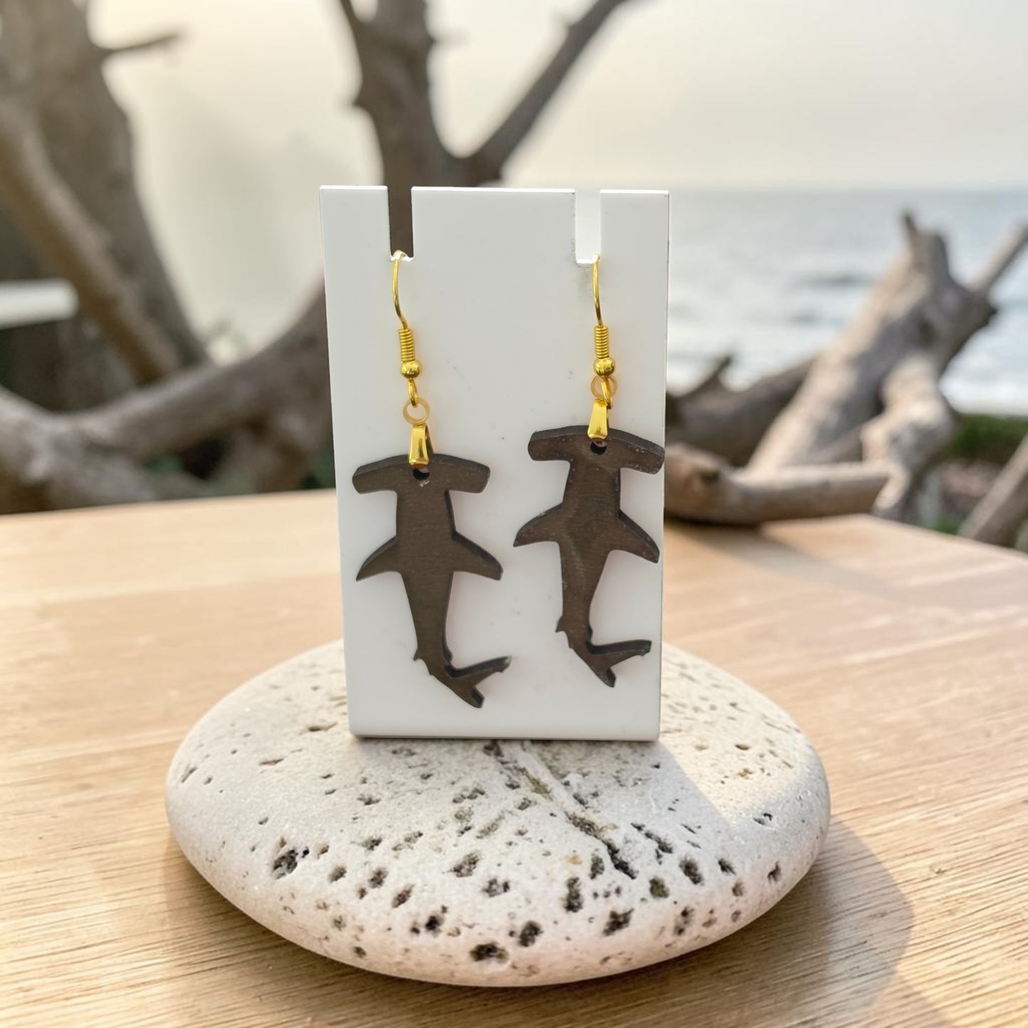 Hammerhead Shark Earrings by Mirabilia Boutique, lasercut from walnut wood, hang on a white display card atop a speckled stone. Driftwood and a blurred ocean view form a coastal backdrop.
