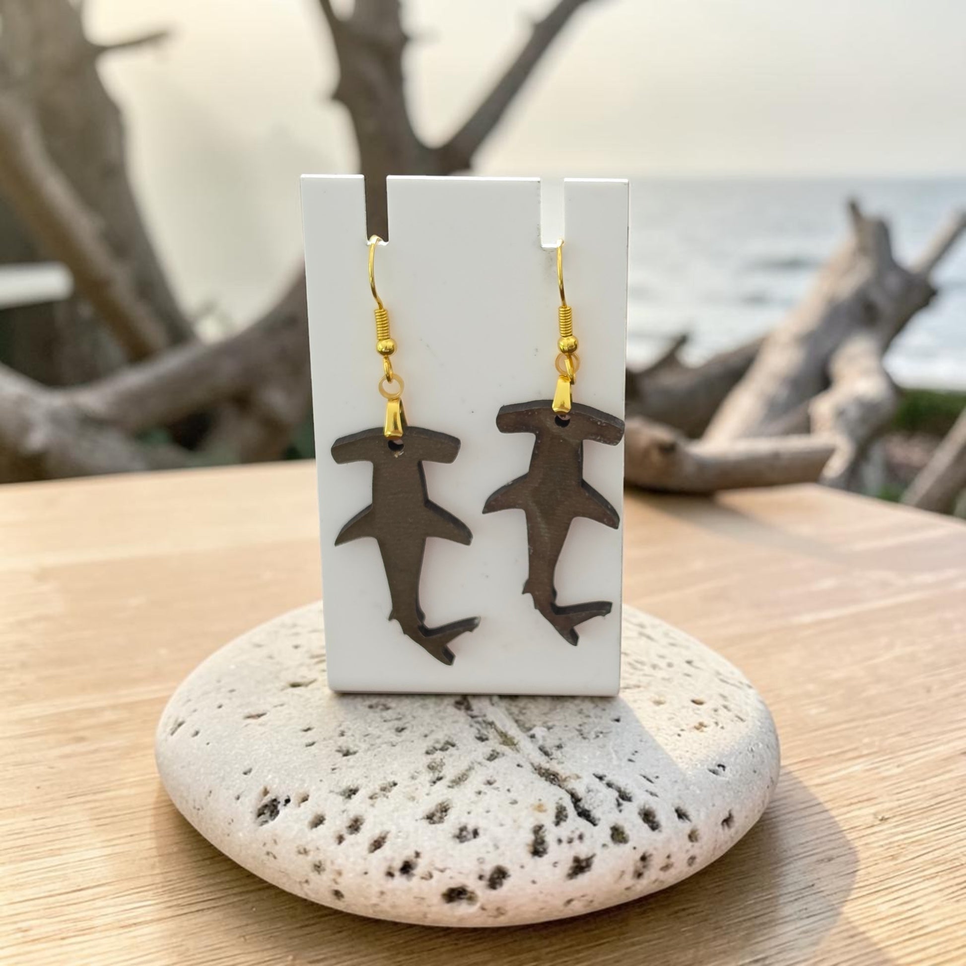 Hammerhead Shark Earrings by Mirabilia Boutique, lasercut from walnut wood, hang on a white display card atop a speckled stone. Driftwood and a blurred ocean view form a coastal backdrop.