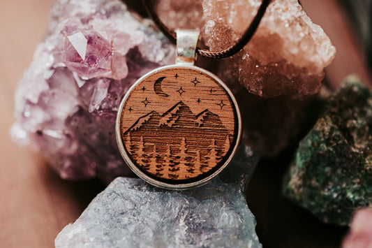 The Engraved Wood Pendant Necklace – Mountain Night Sky Design by Mirabilia Boutique features a round wooden pendant with mountains, trees, and celestial motifs—a dreamy boho accessory for nature lovers.