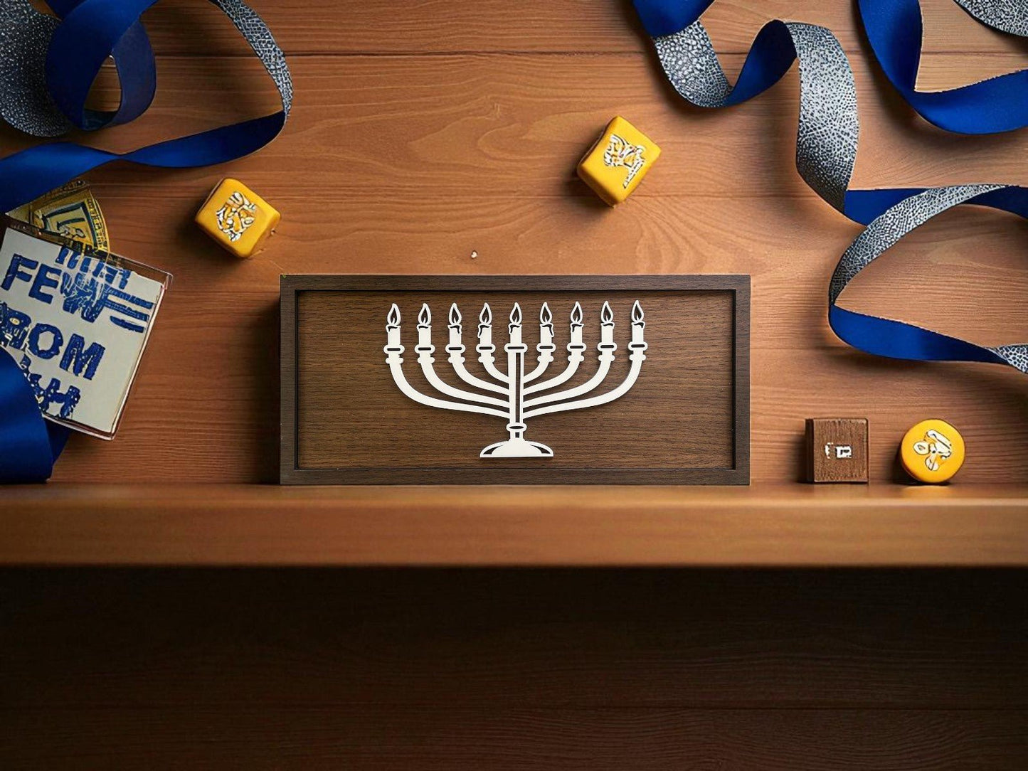 A wooden shelf displays Mirabilia Boutique’s Minimalist Menorah Decor, a 9x4 laser-cut Hanukkah sign, alongside dreidels, blue-and-white ribbon, and a bilingual book to celebrate Hanukkah.