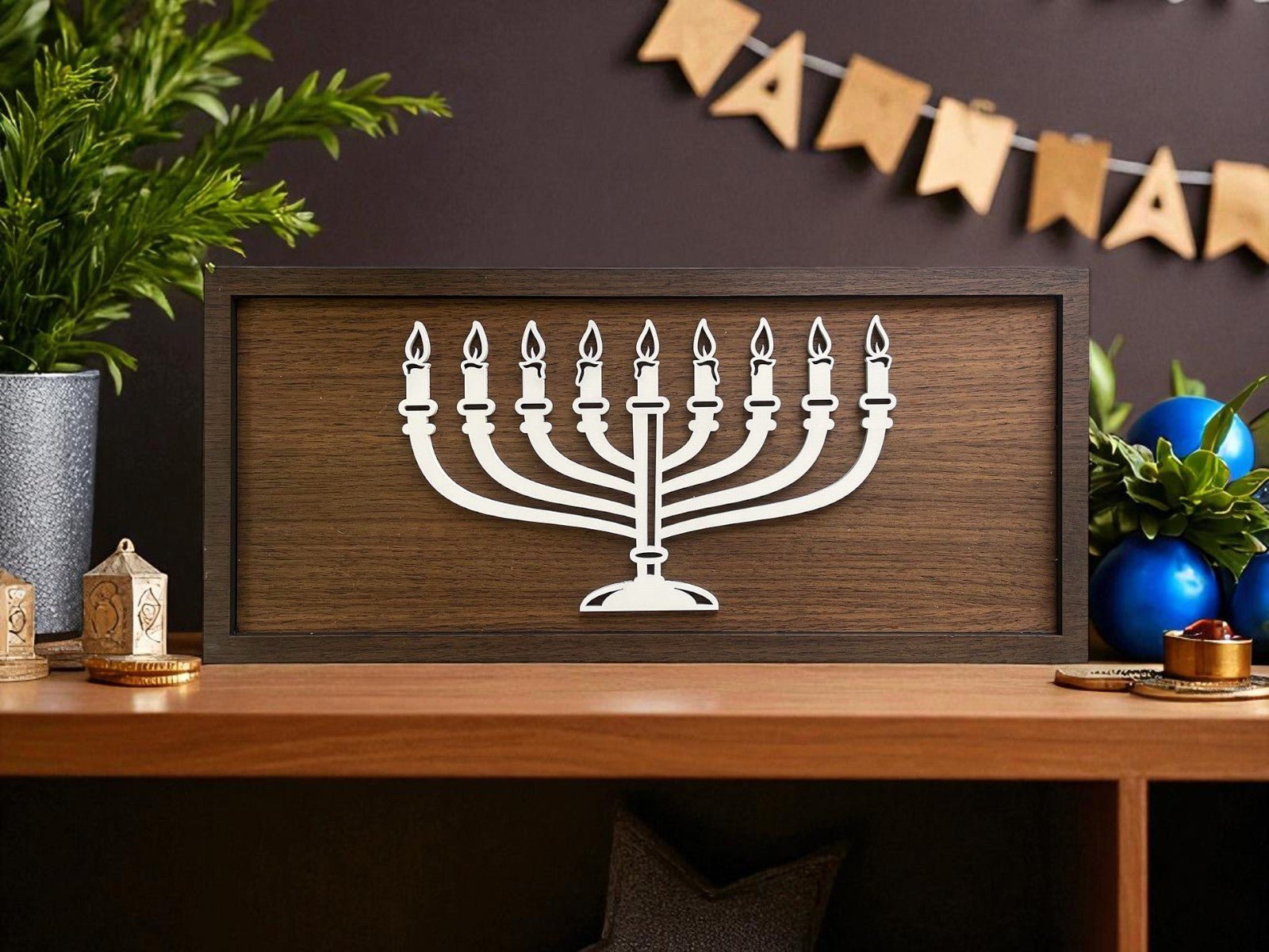 The 18x8 Modern Menorah Sign by Mirabilia Boutique features a white laser-cut menorah with nine lit candles in a wooden frame, displayed on a shelf with plants, blue ornaments, dreidels, and festive Hanukkah decor.
