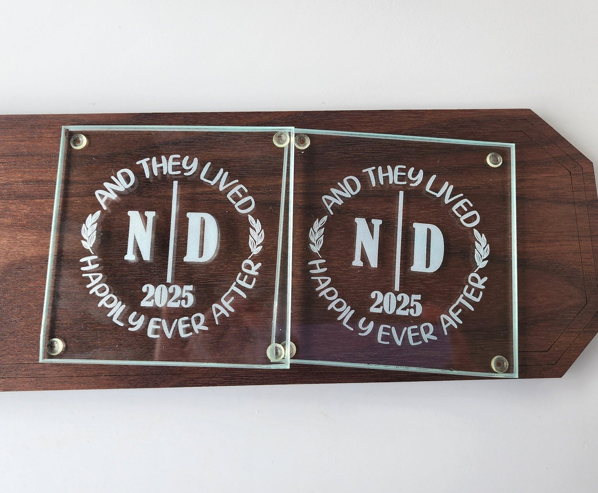 The Personalized Glass Wedding Coasters by Mirabilia Boutique feature elegantly engraved "ND" and "2025" on a wooden plaque with the phrase "And they lived happily ever after," making them an ideal romantic home décor and a perfect personalized wedding gift.