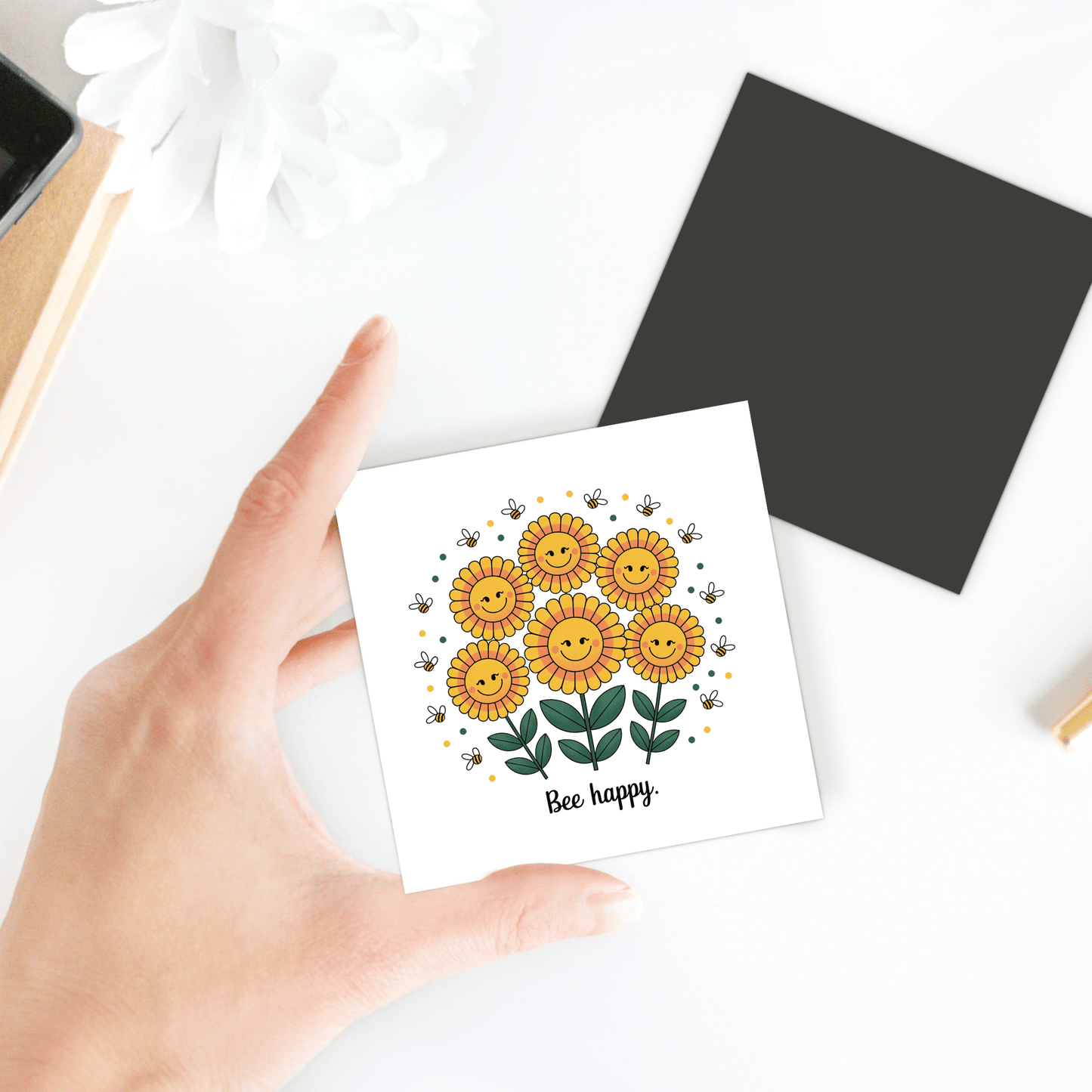 A hand holds the Mirabilia Boutique "Bee Happy Magnet – Cheerful Sunflowers for Daily Joy," featuring smiling sunflowers, small bees, and the words "Bee happy." on a white background. A black square and a white flower are nearby.