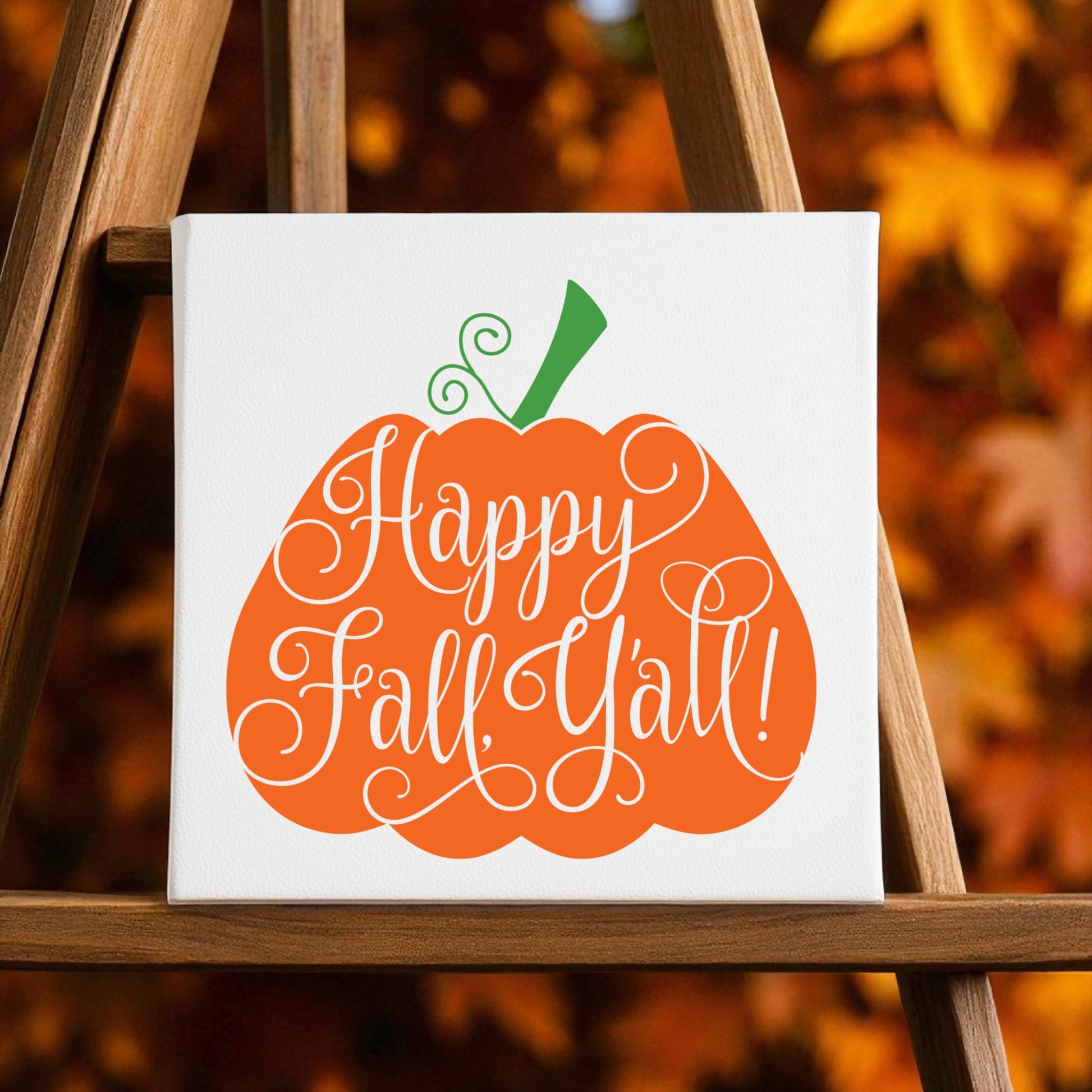 The Happy Fall Y’all Pumpkin Wall Art by Mirabilia Boutique features an orange pumpkin with a curly green stem and "Happy Fall, Y'all!" in white script, set against blurred autumn leaves for cozy southern charm in your fall home decor.