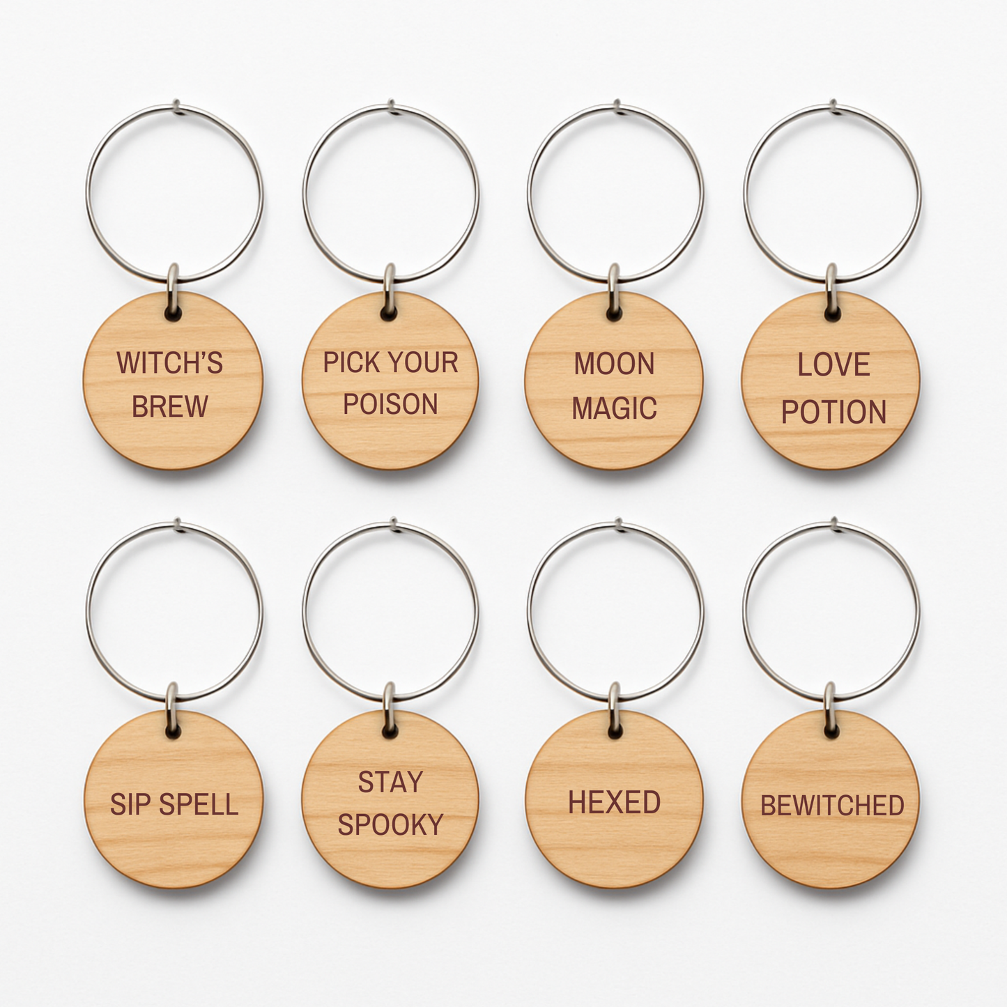The Haunted Harvest Rustic Wine Charms Set by Mirabilia Boutique includes eight wooden keychains with round tags, each laser-engraved with a unique witchy phrase—WITCH'S BREW, PICK YOUR POISON, MOON MAGIC, LOVE POTION, SIP SPELL, STAY SPOOKY, HEXED, and BEWITCHED. Perfect for using as spooky wine glass markers or Halloween wine charms at your next magically rustic gathering.