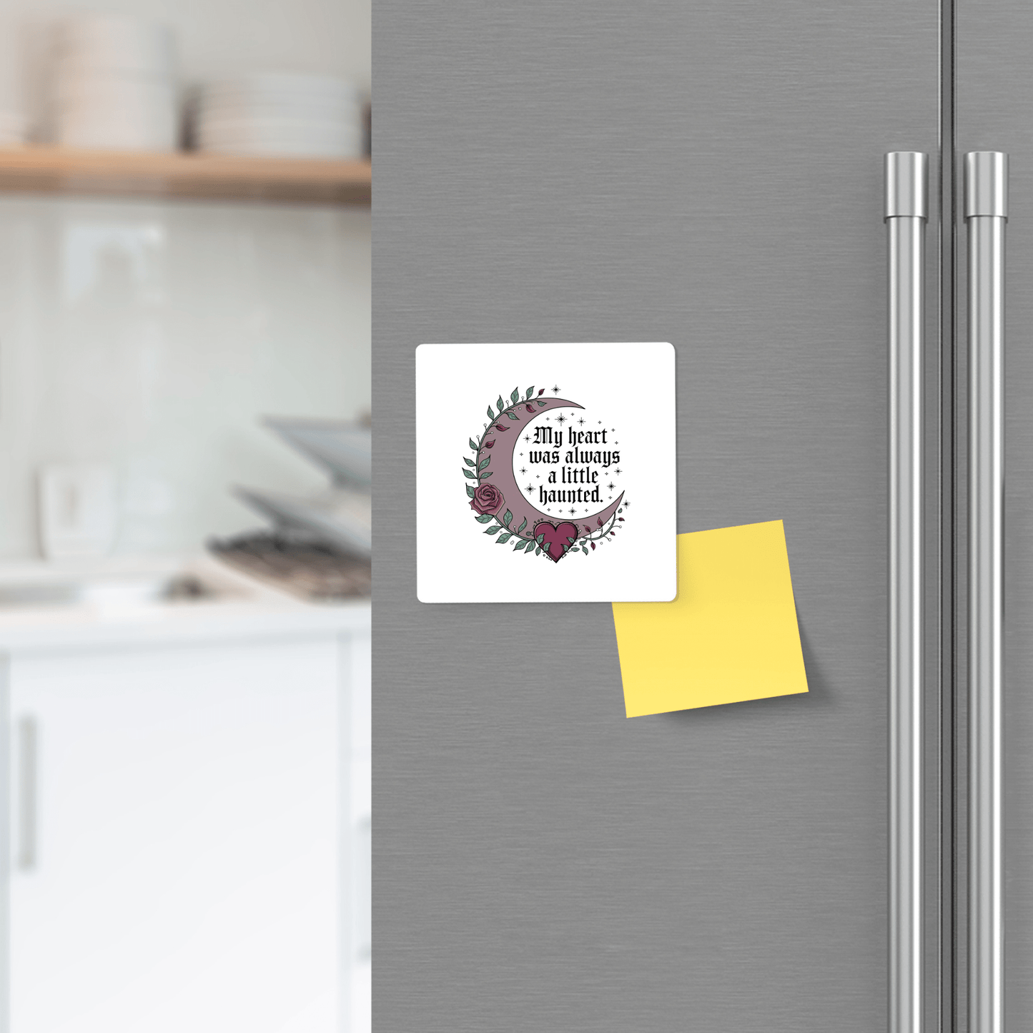 A Mirabilia Boutique Gothic Fridge Magnet featuring a floral crescent moon and "My heart was always a little haunted" decorates a refrigerator next to a blank yellow sticky note, with a kitchen counter in the background.