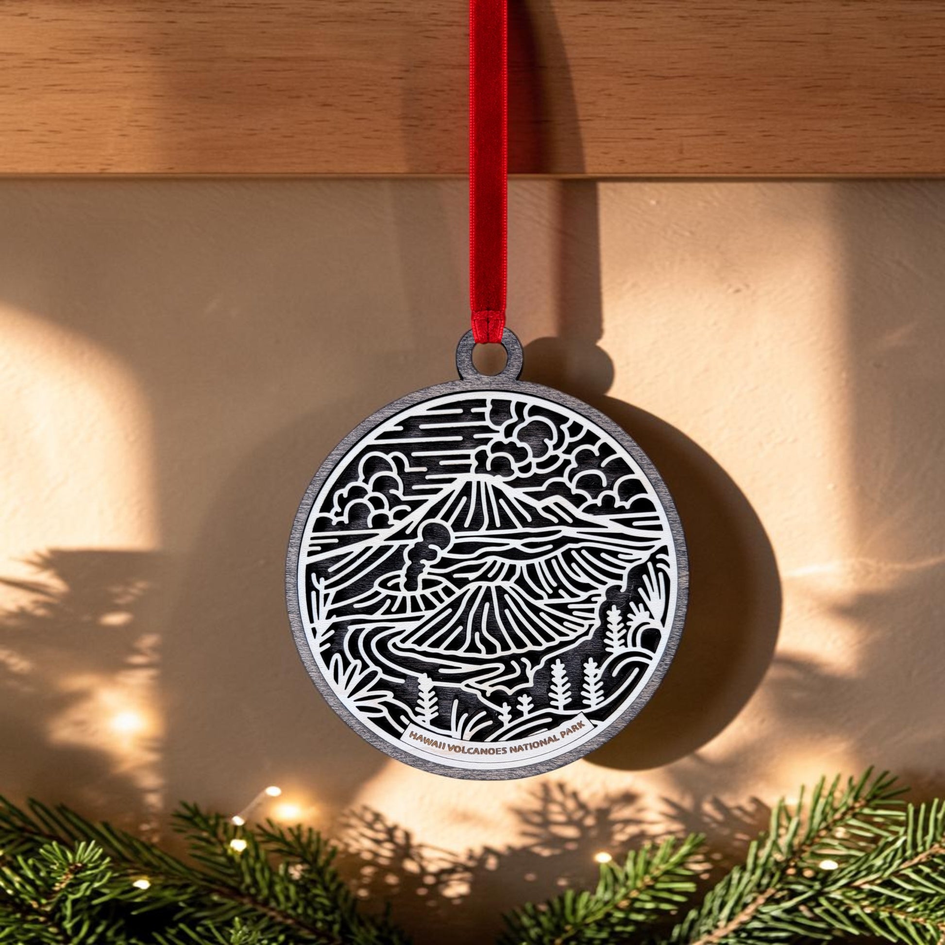 The Hawaiʻi Volcanoes National Park Line Art Christmas Ornament by Mirabilia Boutique features a round wooden design adorned with a black and white volcanic landscape. Hanging from a red ribbon, the ornament showcases sun rays, mountains, and plants inspired by the stunning scenery of Hawaiʻi Volcanoes National Park. A garland of greenery beneath adds a touch of natural elegance.
