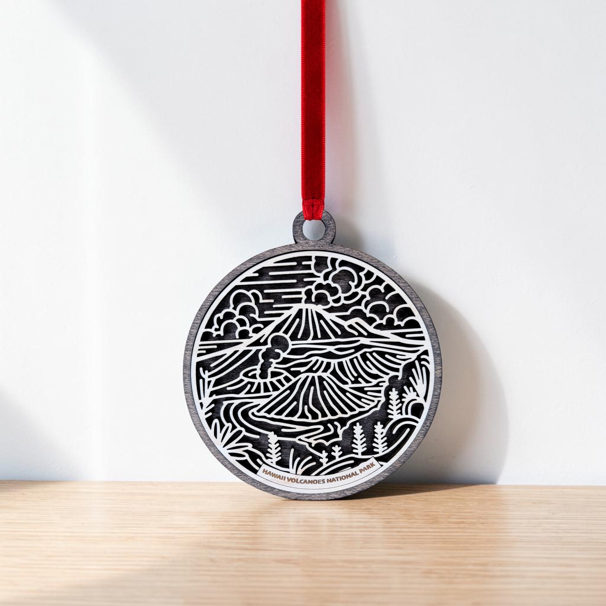 A round handcrafted wooden ornament from Mirabilia Boutique, the Hawaiʻi Volcanoes National Park Line Art Christmas Ornament features a black and white volcanic landscape design. Hanging by a red ribbon against a white wall and resting on a wooden surface, this beautiful keepsake is perfect for any nature lover.
