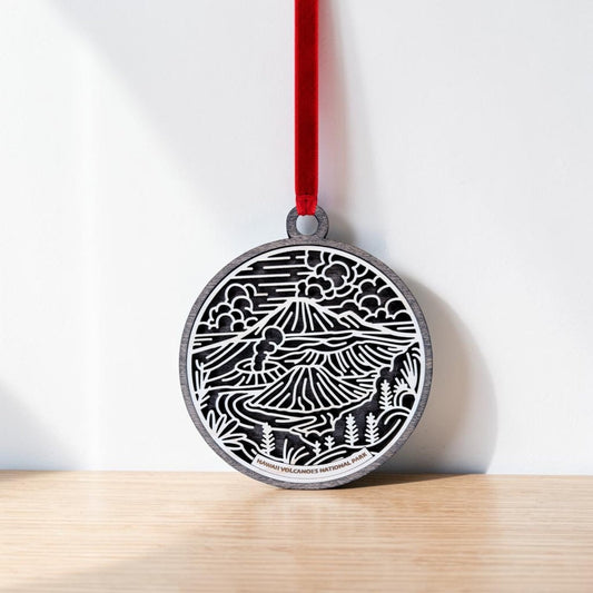 A round handcrafted wooden ornament from Mirabilia Boutique, the Hawaiʻi Volcanoes National Park Line Art Christmas Ornament features a black and white volcanic landscape design. Hanging by a red ribbon against a white wall and resting on a wooden surface, this beautiful keepsake is perfect for any nature lover.