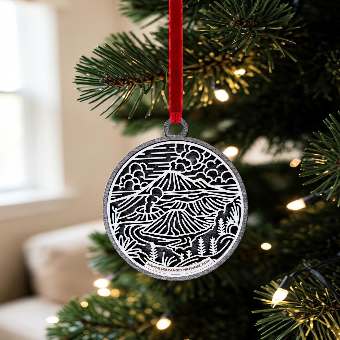 A handcrafted Hawaiʻi Volcanoes National Park Line Art Christmas Ornament by Mirabilia Boutique, showcasing a scenic black and white mountain and forest design, hangs on a Christmas tree branch adorned with white lights and a red ribbon.