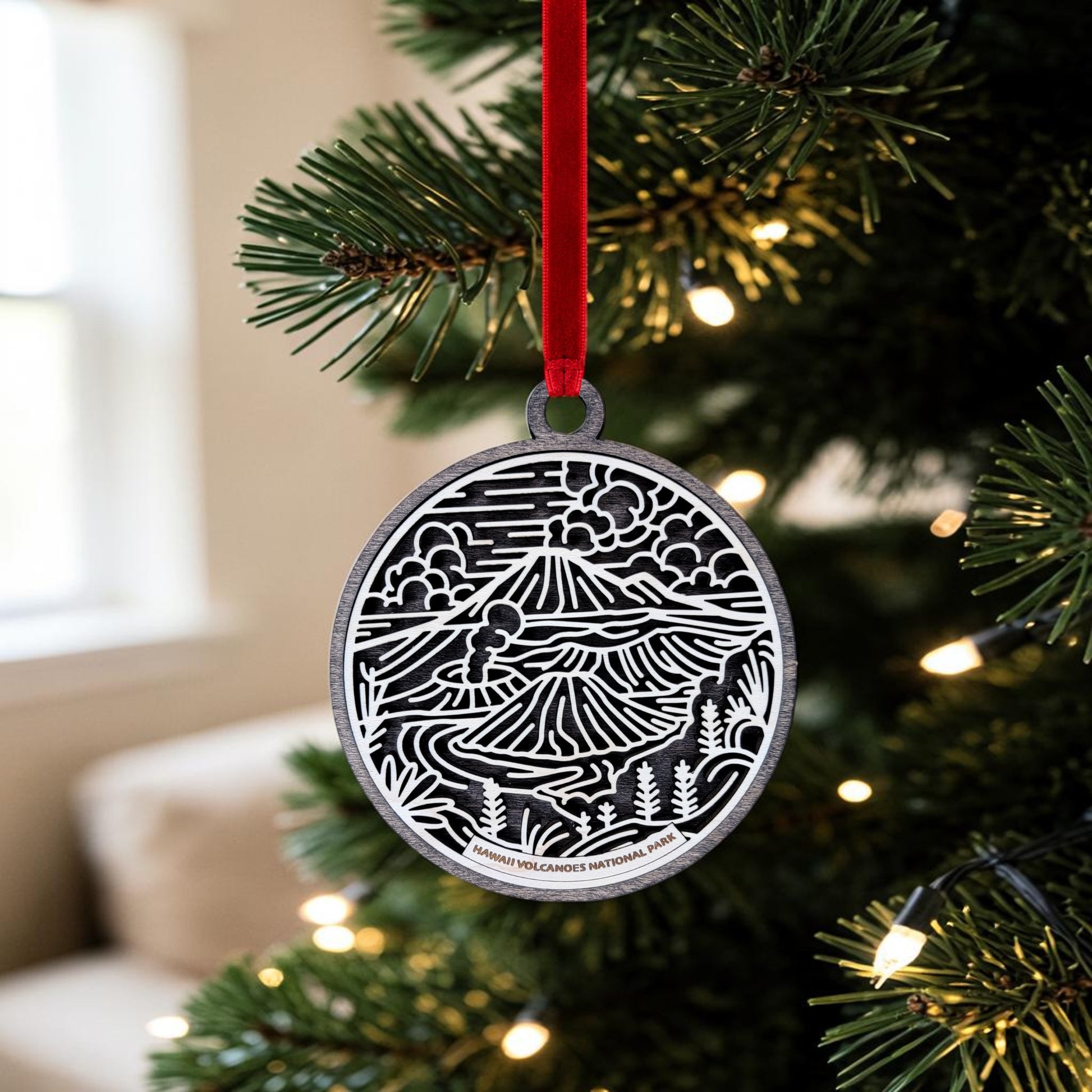 A handcrafted Hawaiʻi Volcanoes National Park Line Art Christmas Ornament by Mirabilia Boutique, showcasing a scenic black and white mountain and forest design, hangs on a Christmas tree branch adorned with white lights and a red ribbon.